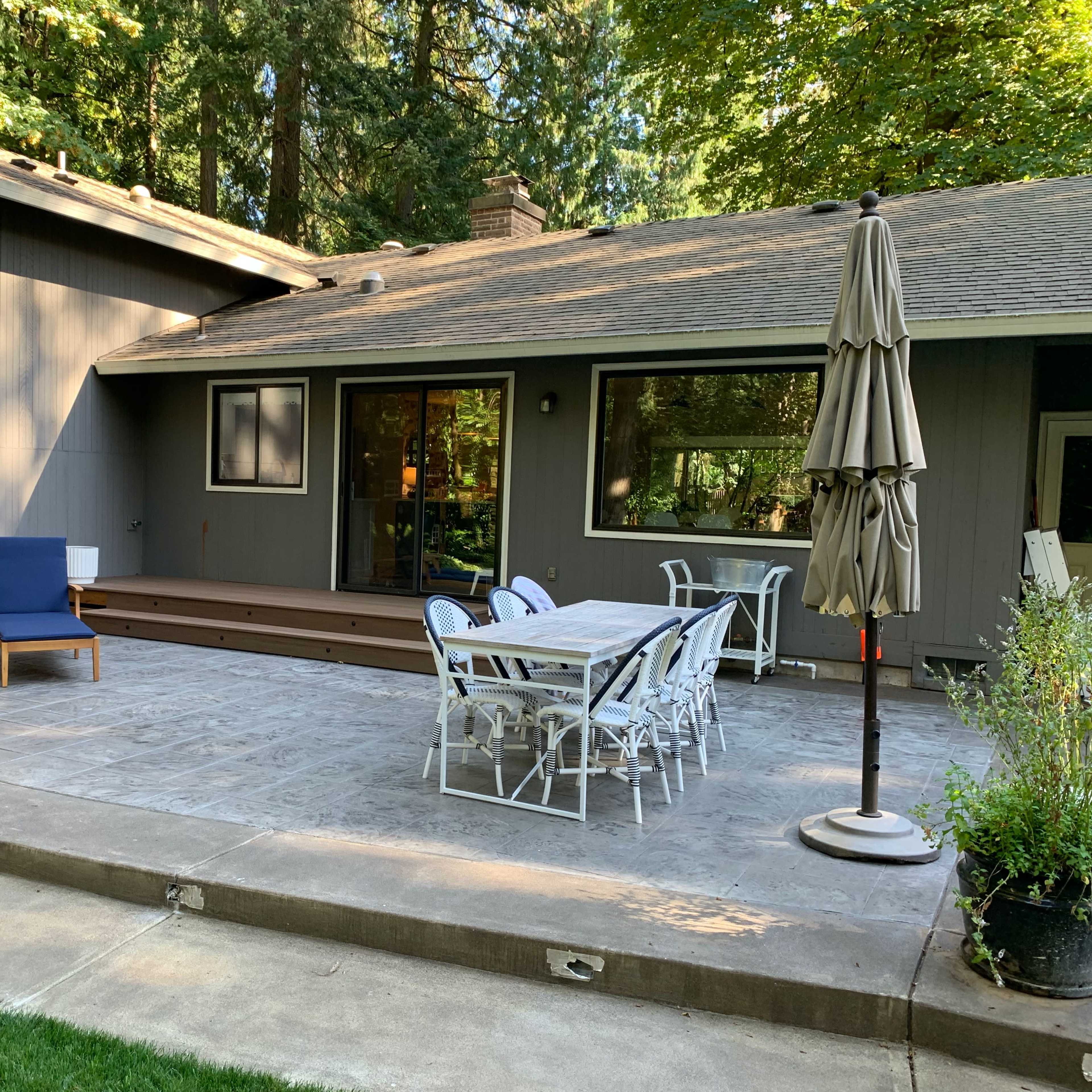 A patio area with a dining table and chairs, two lounge chairs, and an umbrella in front of a house surrounded by trees.