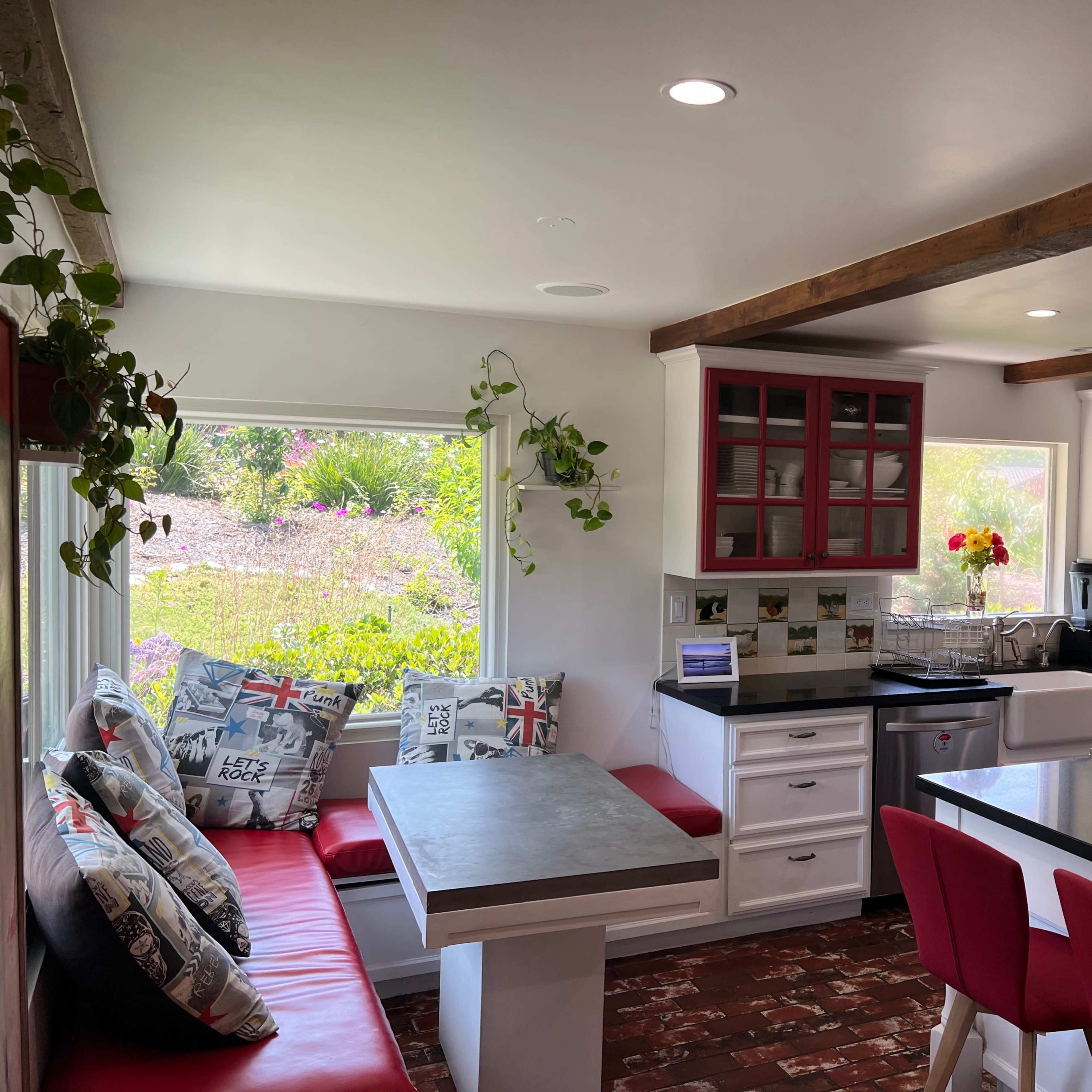 A modern kitchen features a red and white color scheme with a dining area, a central island, and large windows overlooking greenery.