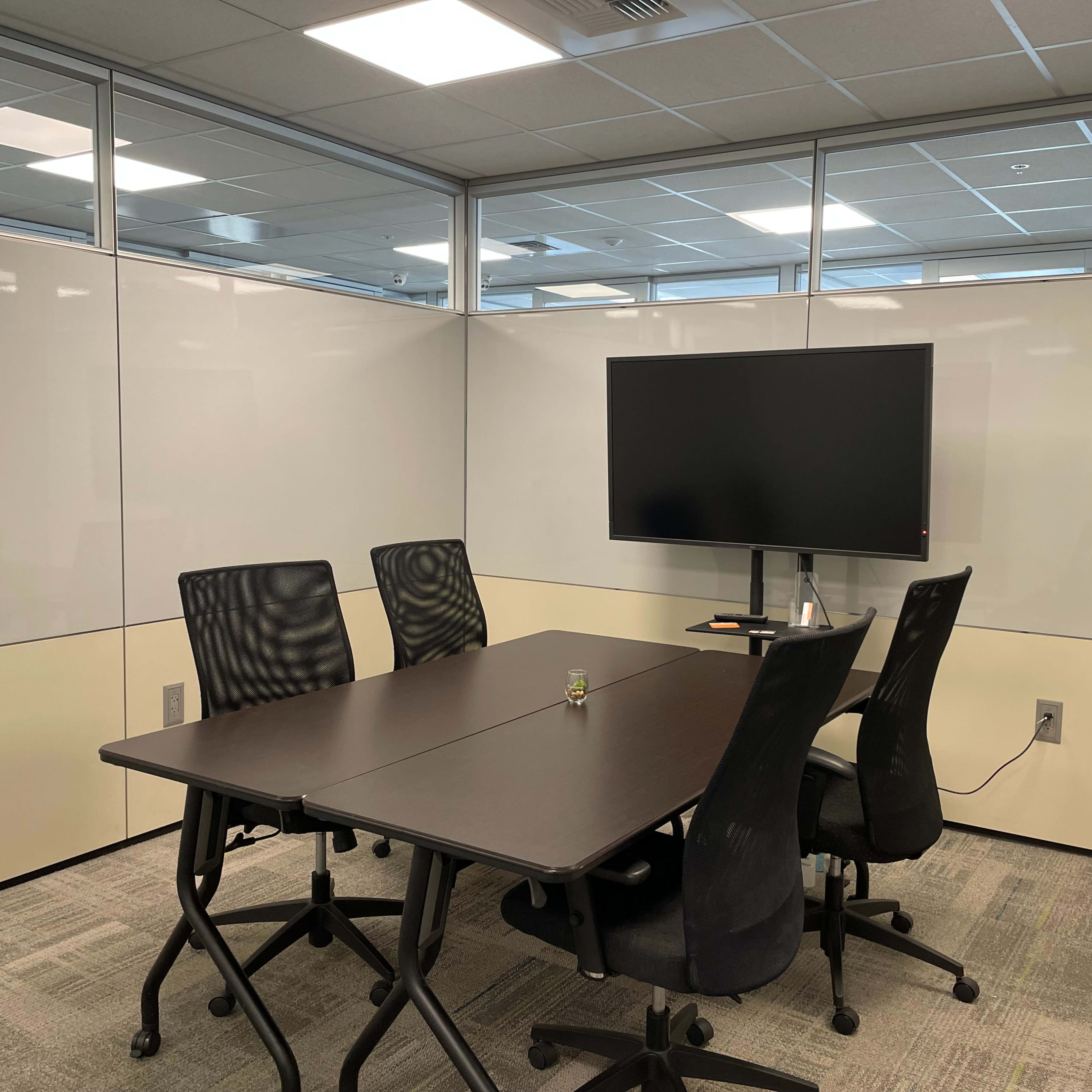 A small conference room with a table, four chairs, and a wall-mounted monitor.