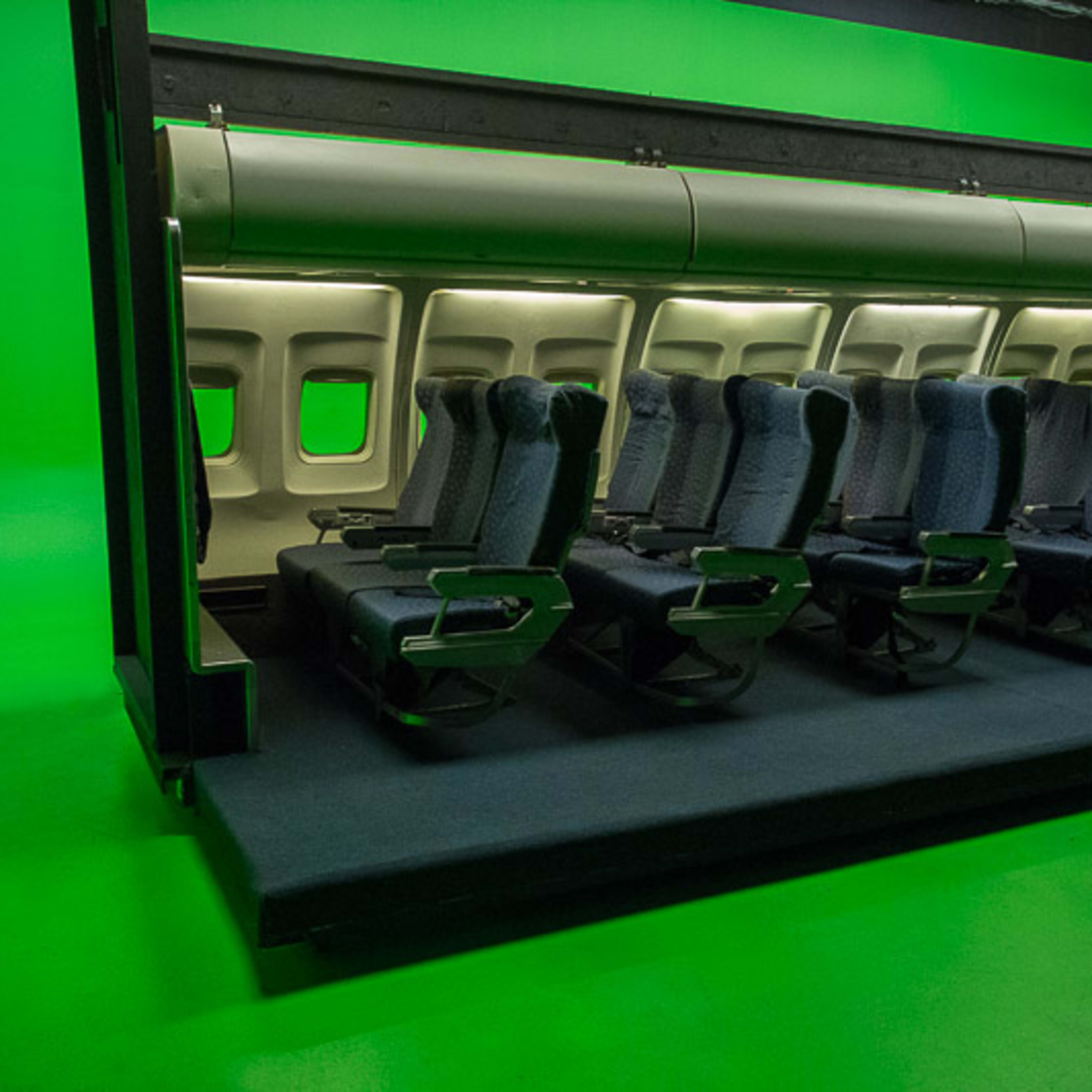 A row of airplane seats is positioned in front of a green backdrop in a film studio.
