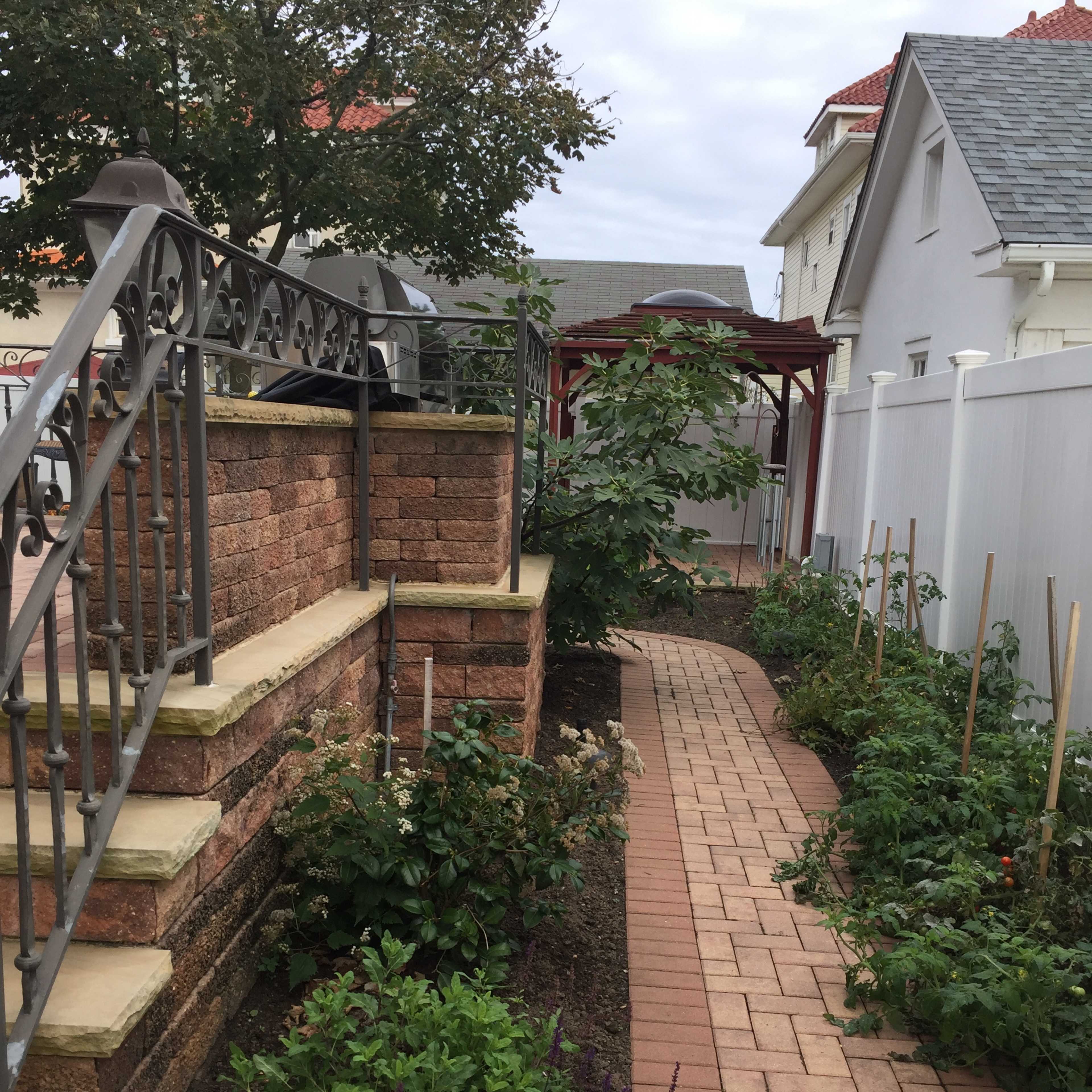 A brick walkway bordered by garden beds leads past stone stairs and residential buildings under an overcast sky.