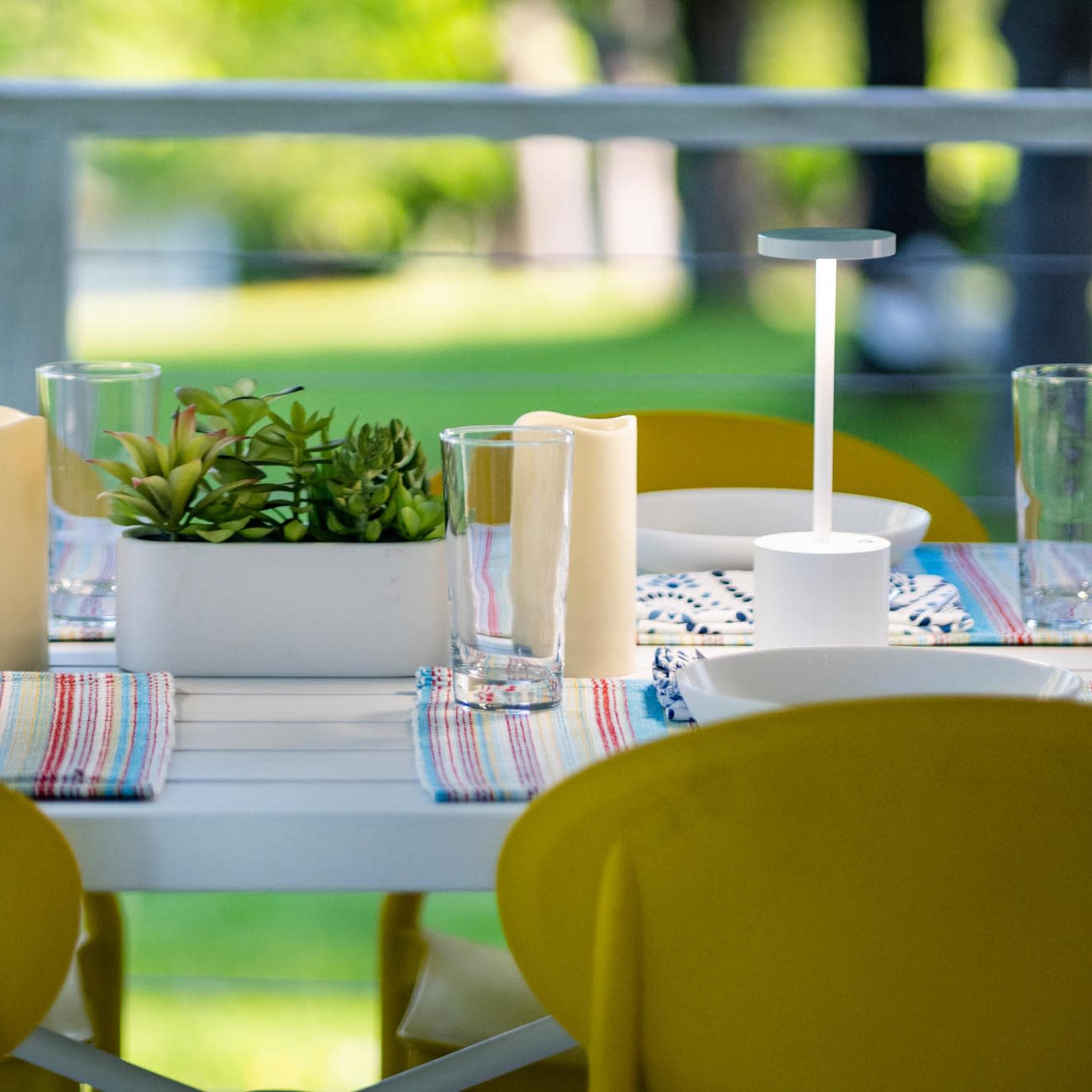 A dining table is set with yellow chairs, white dishes, glasses, and decorative candles, along with a potted plant centerpiece on a striped tablecloth.