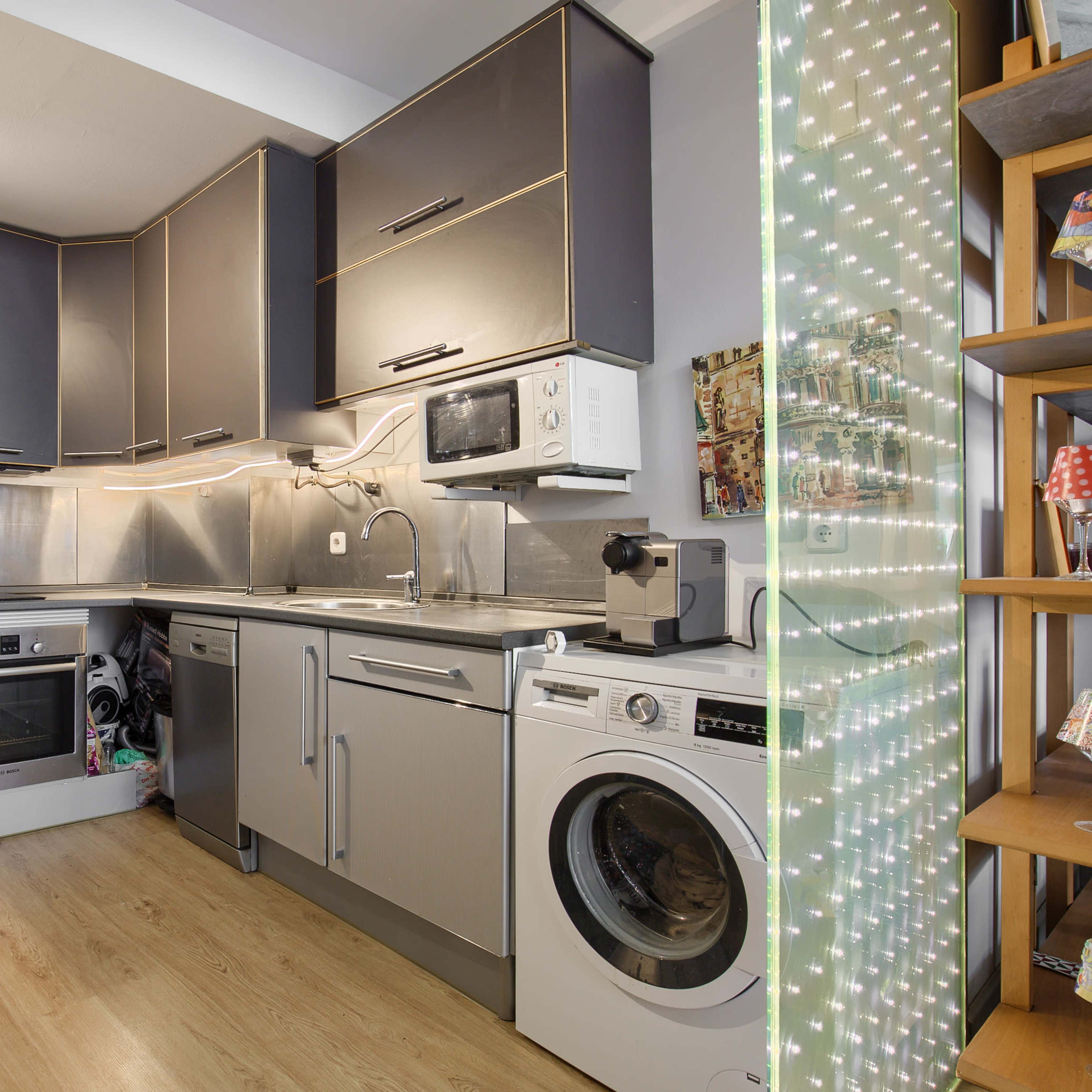 A compact kitchen featuring modern appliances, including a refrigerator, oven, microwave, and washing machine, with a lighted decorative panel on the right.