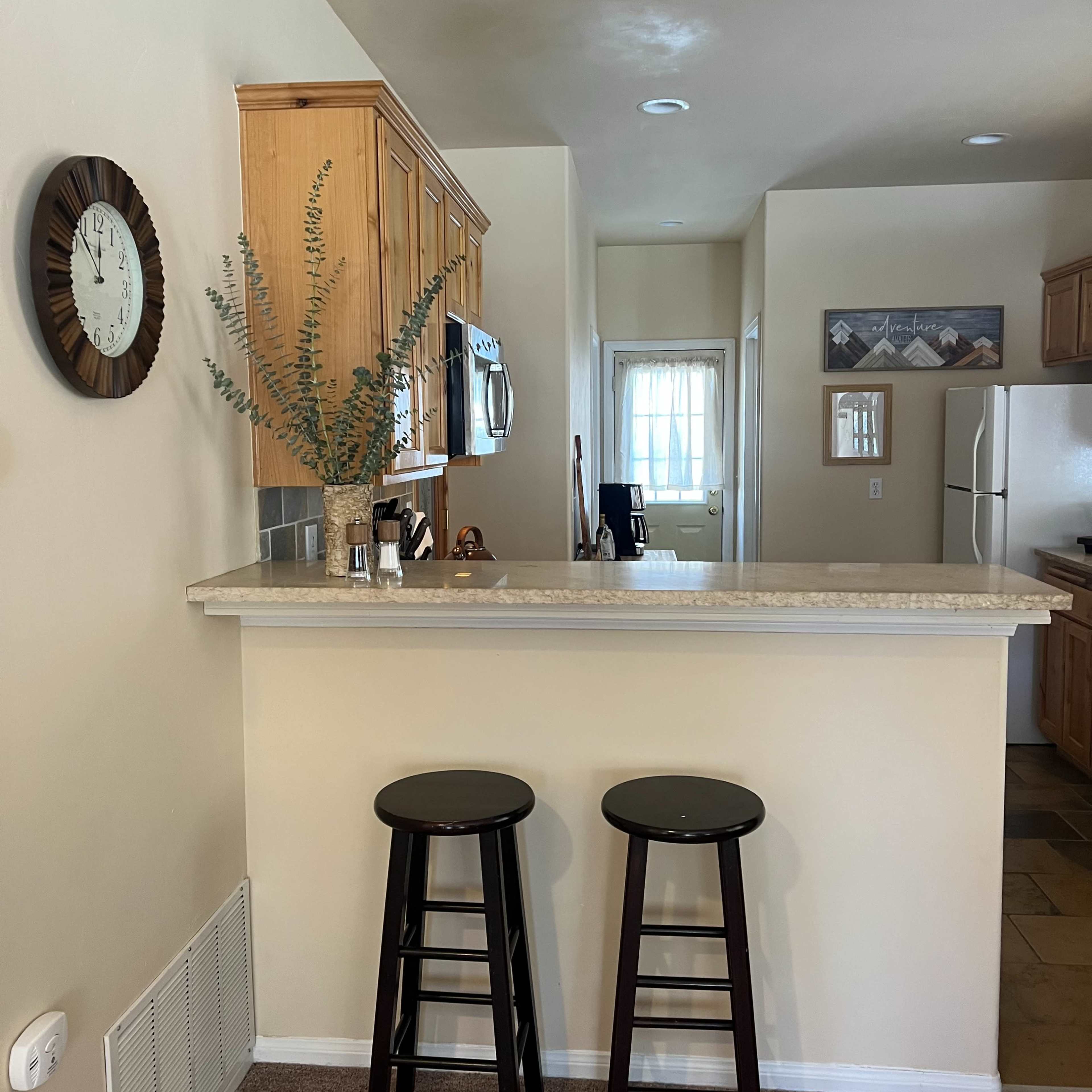 A kitchen area with a counter, two black stools, wooden cabinets, and a clock on the wall.