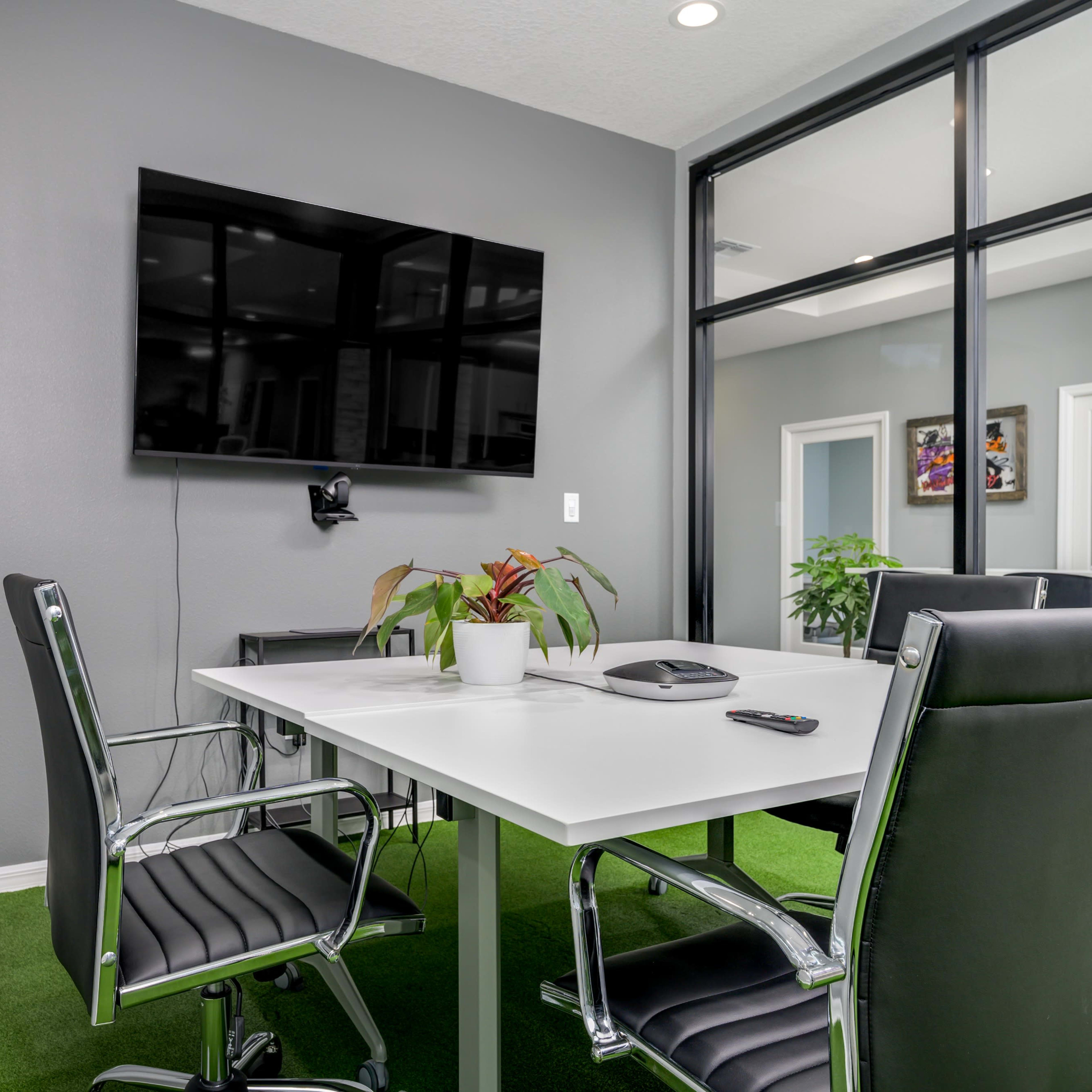 A conference room features a modern table with four black chairs, a wall-mounted TV, and potted plants, all set against a gray backdrop.