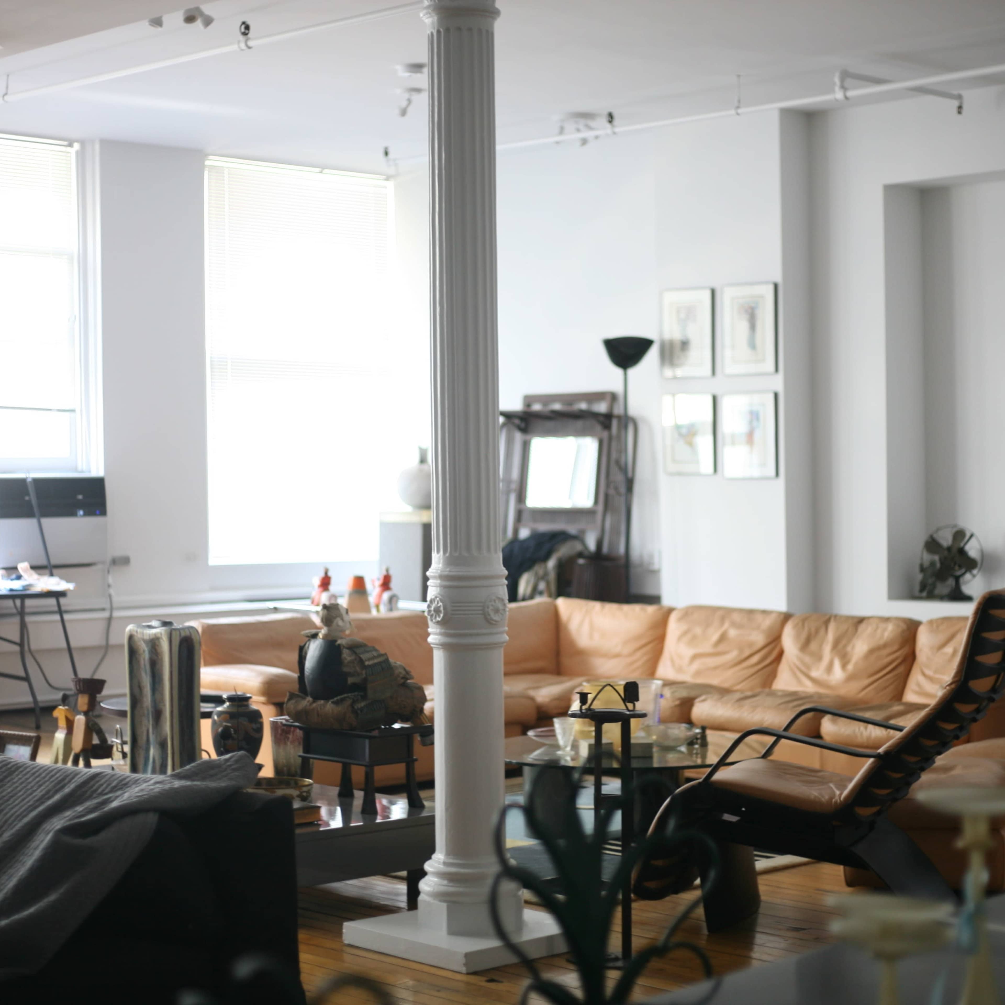 A spacious interior with a tan leather sectional sofa, wooden flooring, tall windows, and various furnishings including a desk and decorative items.