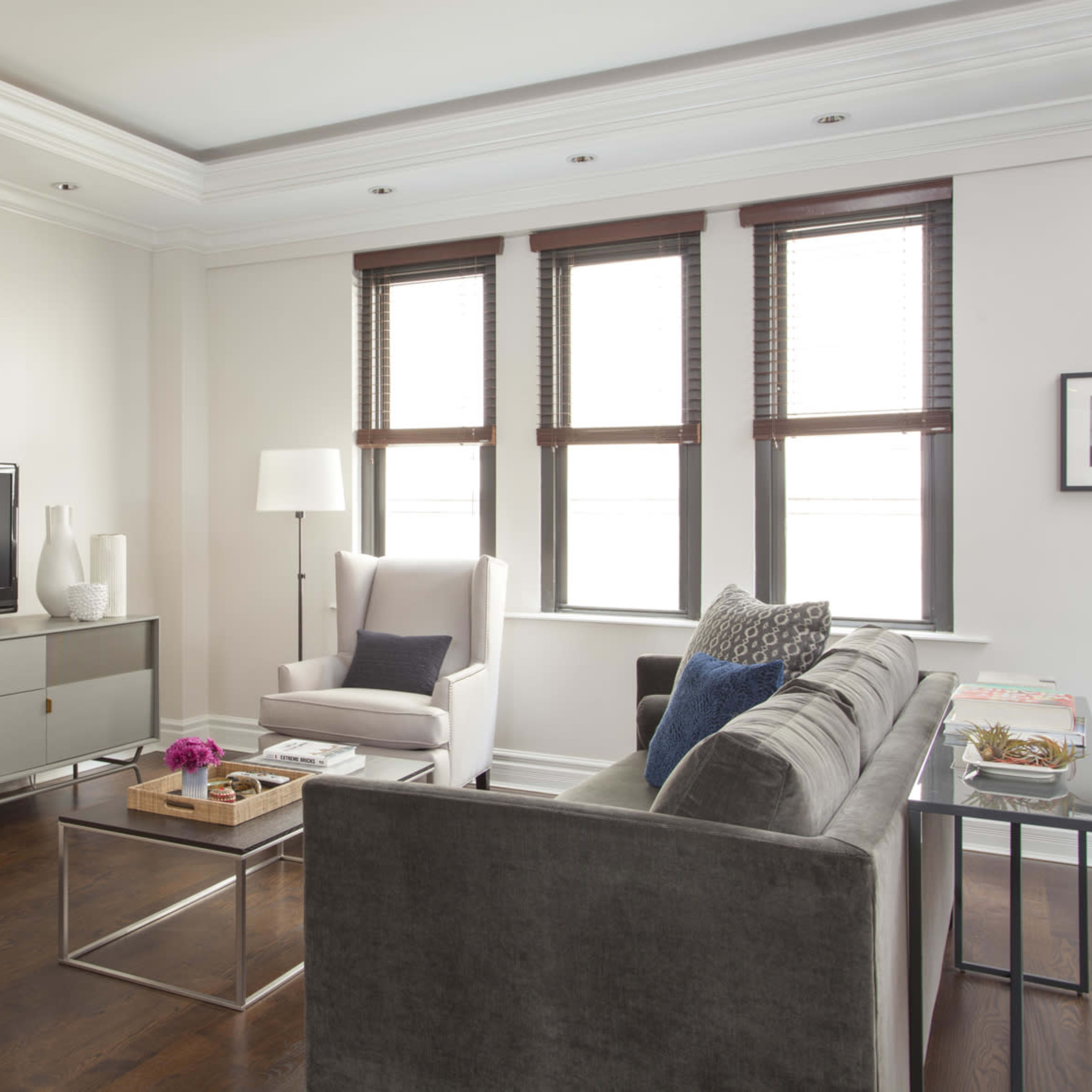 A modern living room features a gray sofa, two armchairs, a television on a media console, and three windows with blinds, illuminated by natural light.