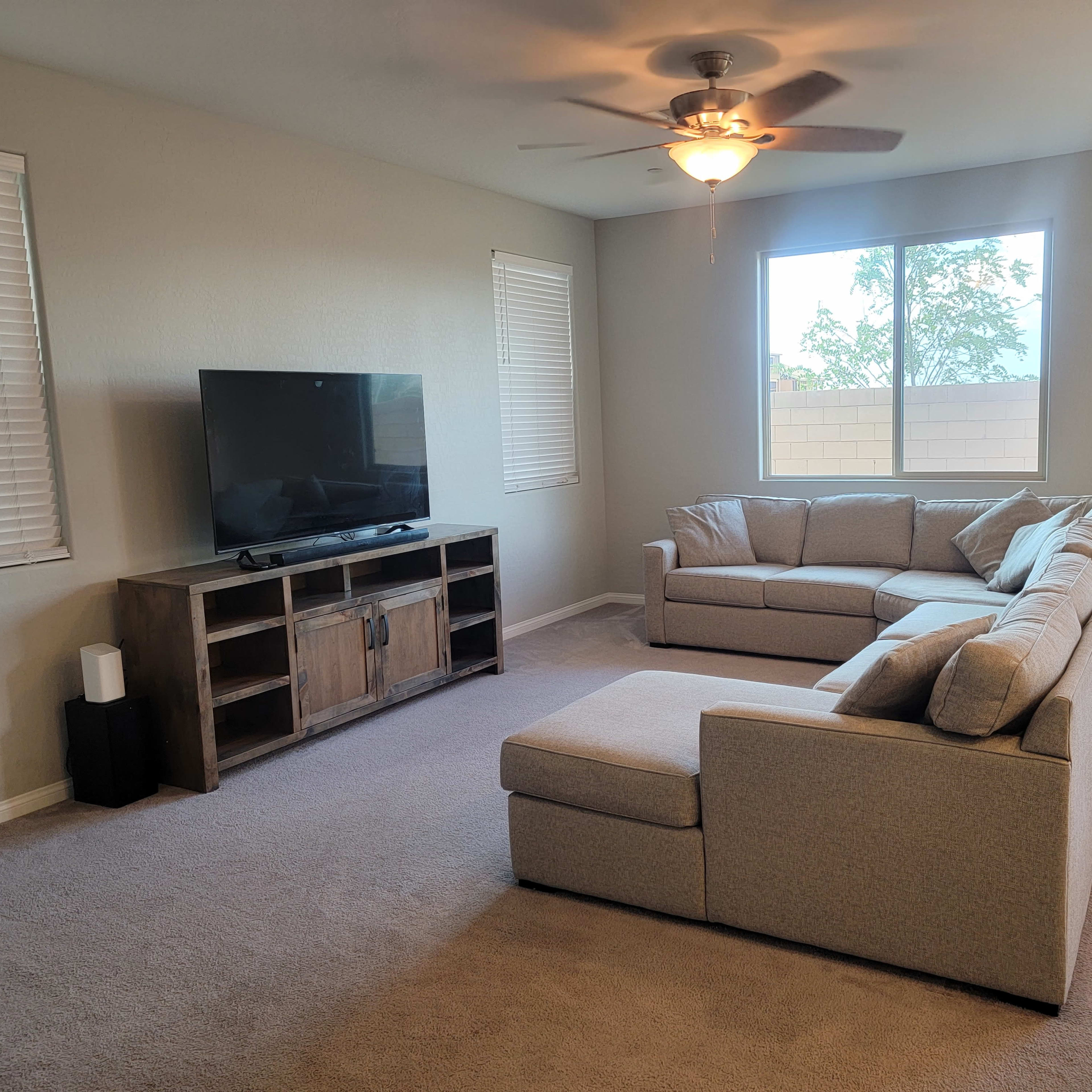 A spacious living room features a large sectional sofa, a television on a wooden stand, and windows allowing natural light to enter.