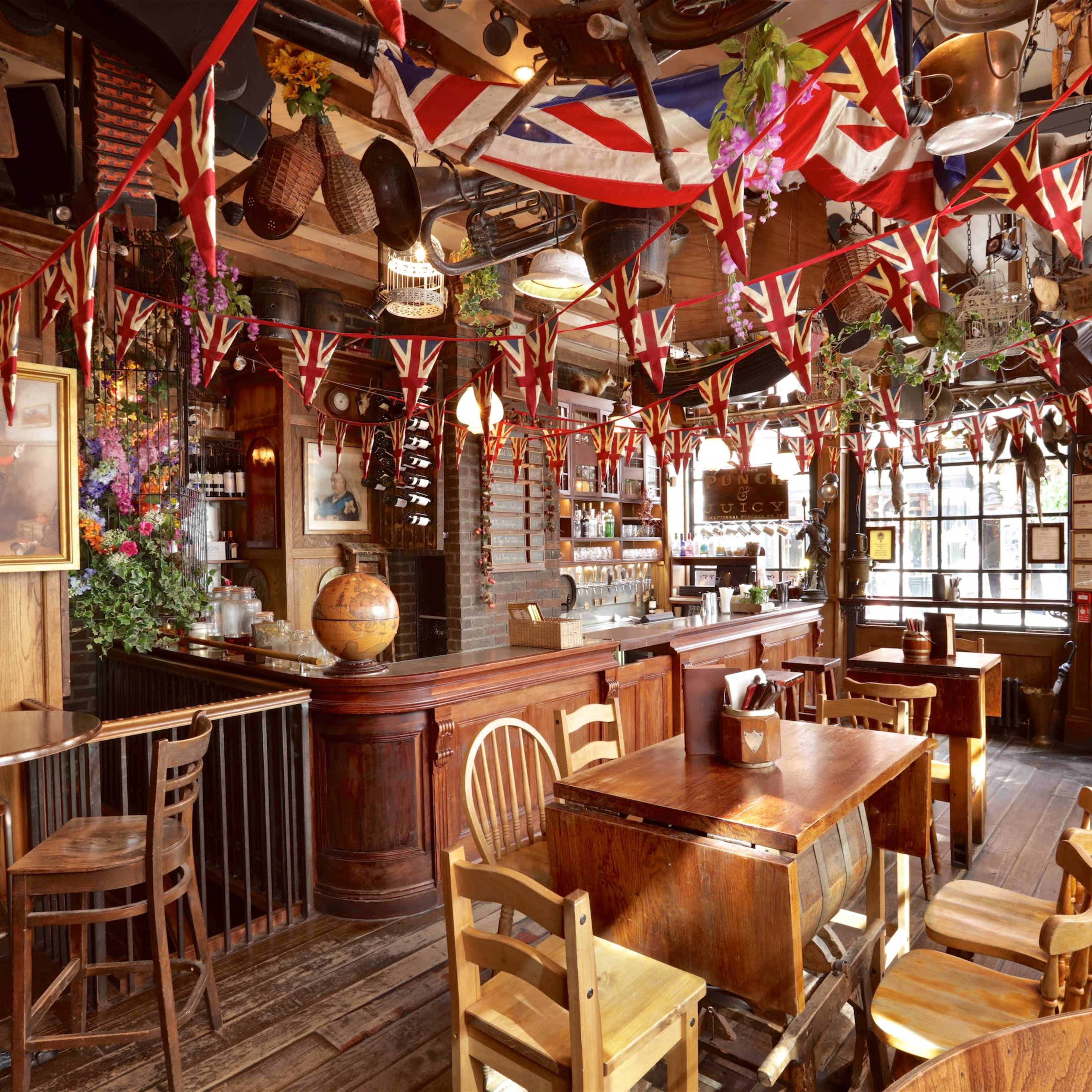 A traditional pub interior features wooden furnishings, a bar with a variety of bottles, and walls adorned with British flags and decorative items.