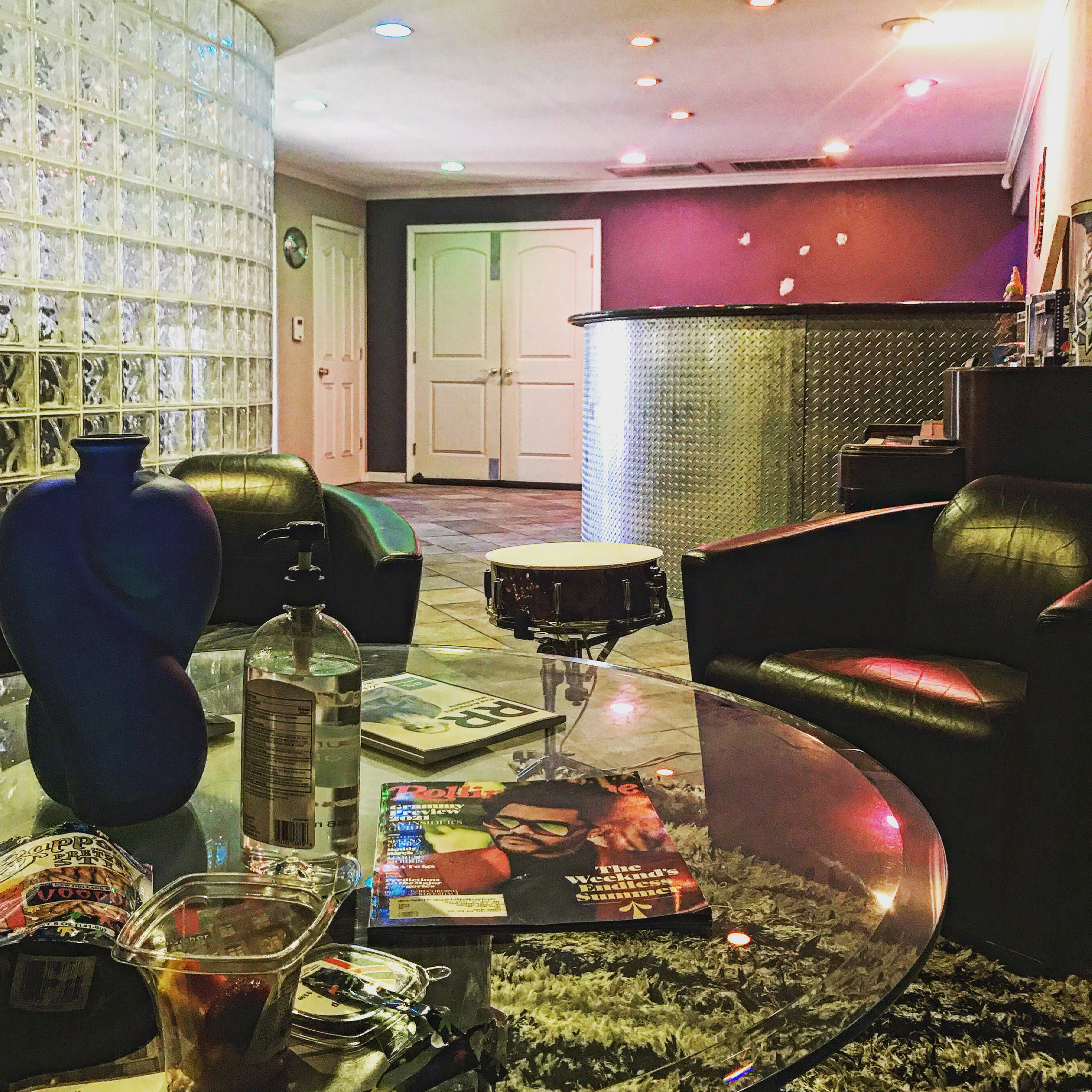 The scene shows a modern interior featuring a glass block wall, a circular bar area, and a coffee table with drinks and magazines in a lounge setting.