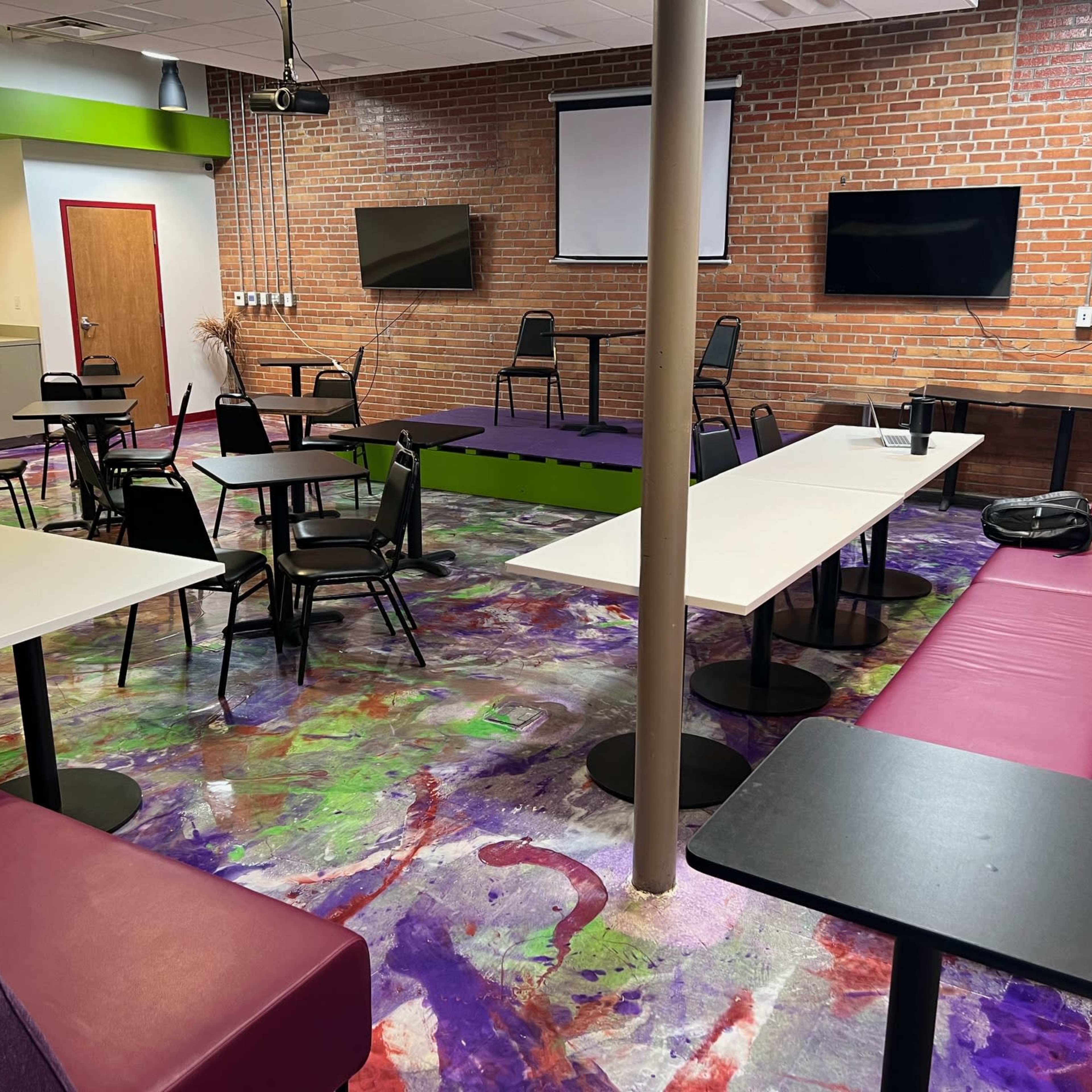 A room with a colorful floor, featuring tables and chairs arranged for seating, a small stage area, and two wall-mounted televisions.