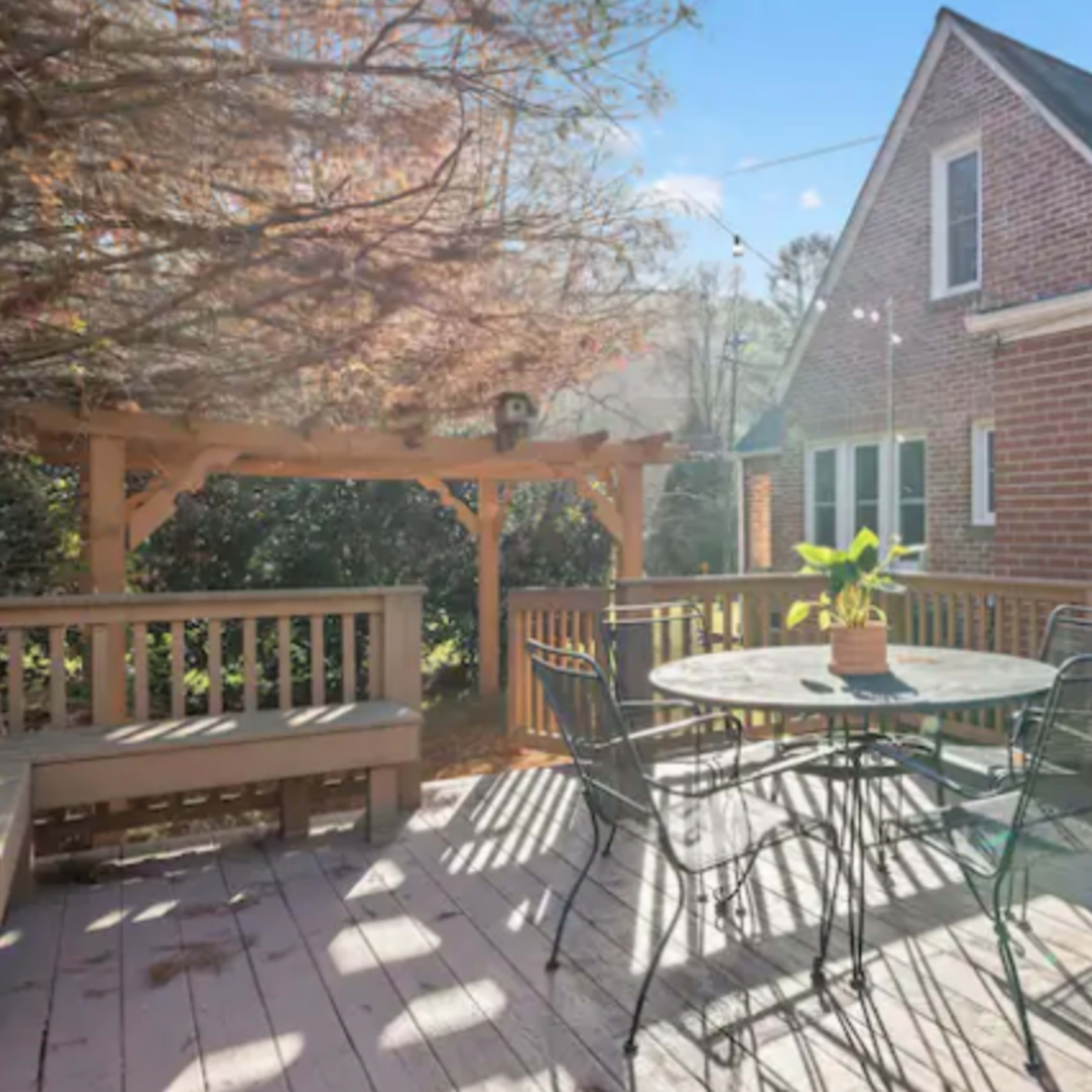 A wooden deck with a circular table and chairs, surrounded by a brick house and a pergola.