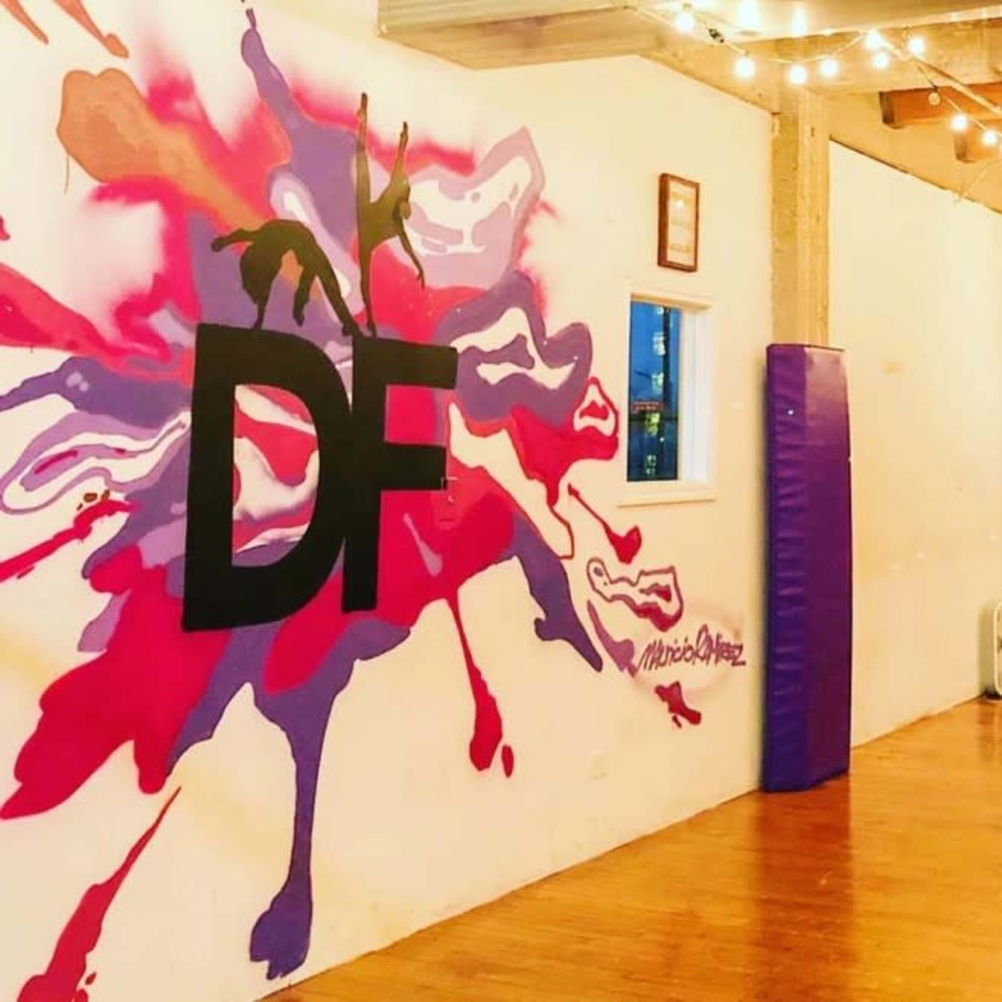 An indoor dance studio with a colorful mural on the wall, wooden flooring, and large windows illuminated by string lights.