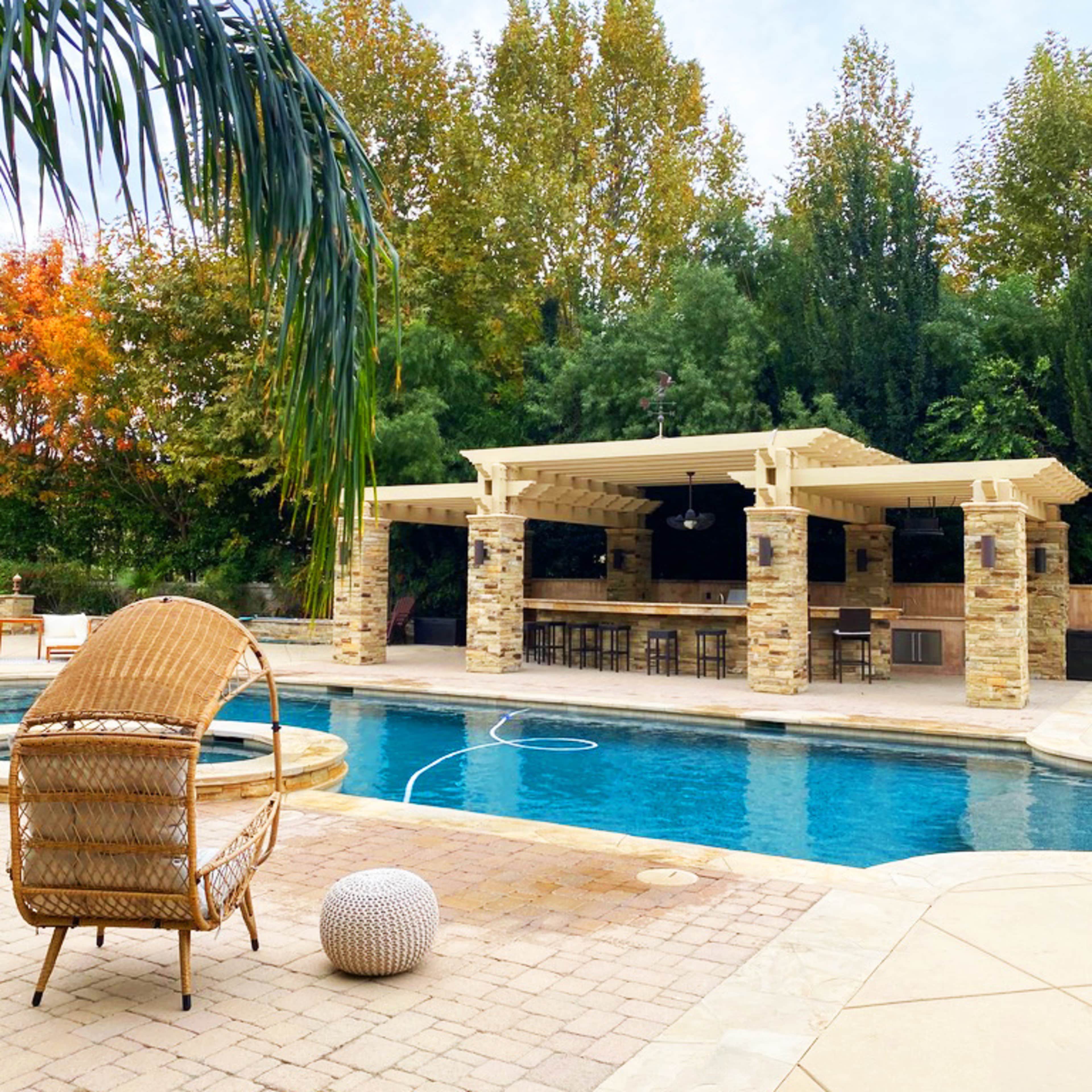 A stone patio surrounds a swimming pool, with a covered outdoor kitchen and seating area visible in the background amid trees displaying autumn colors.