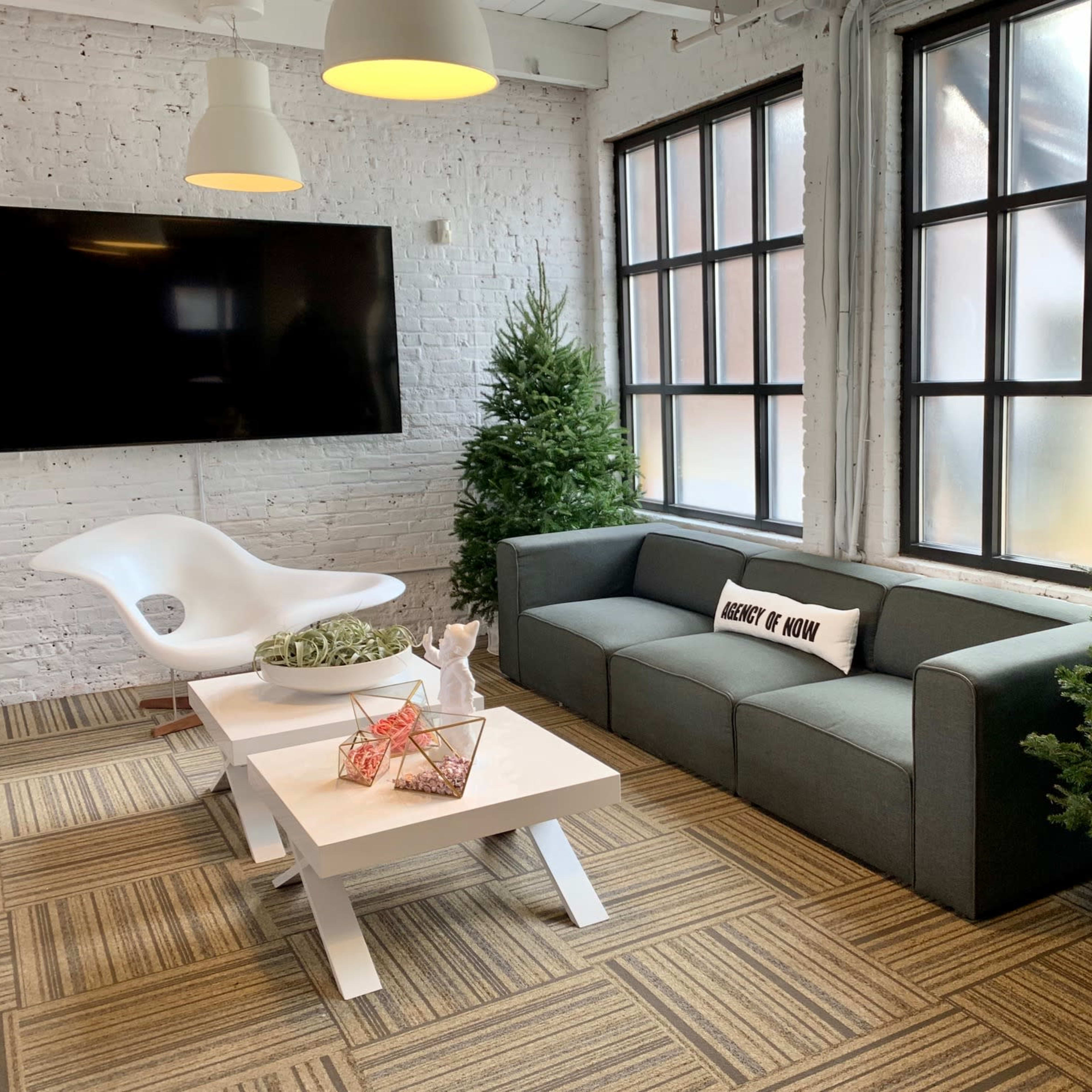 A modern lounge area with a gray sofa, a white coffee table, two small Christmas trees, and a large window with multiple panes.