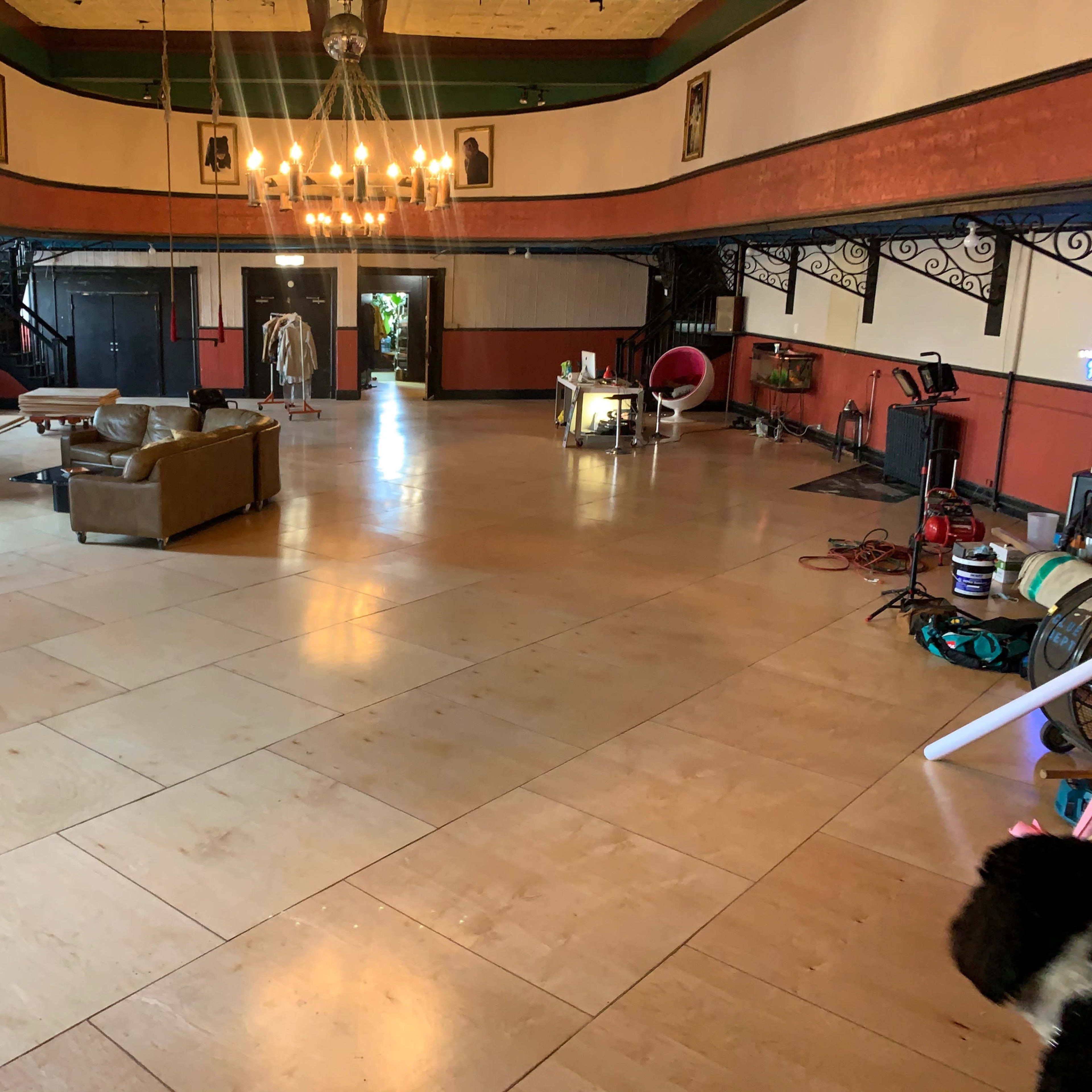 A spacious indoor area with wooden flooring, featuring a few pieces of furniture, including a brown couch and a table, under a chandelier.