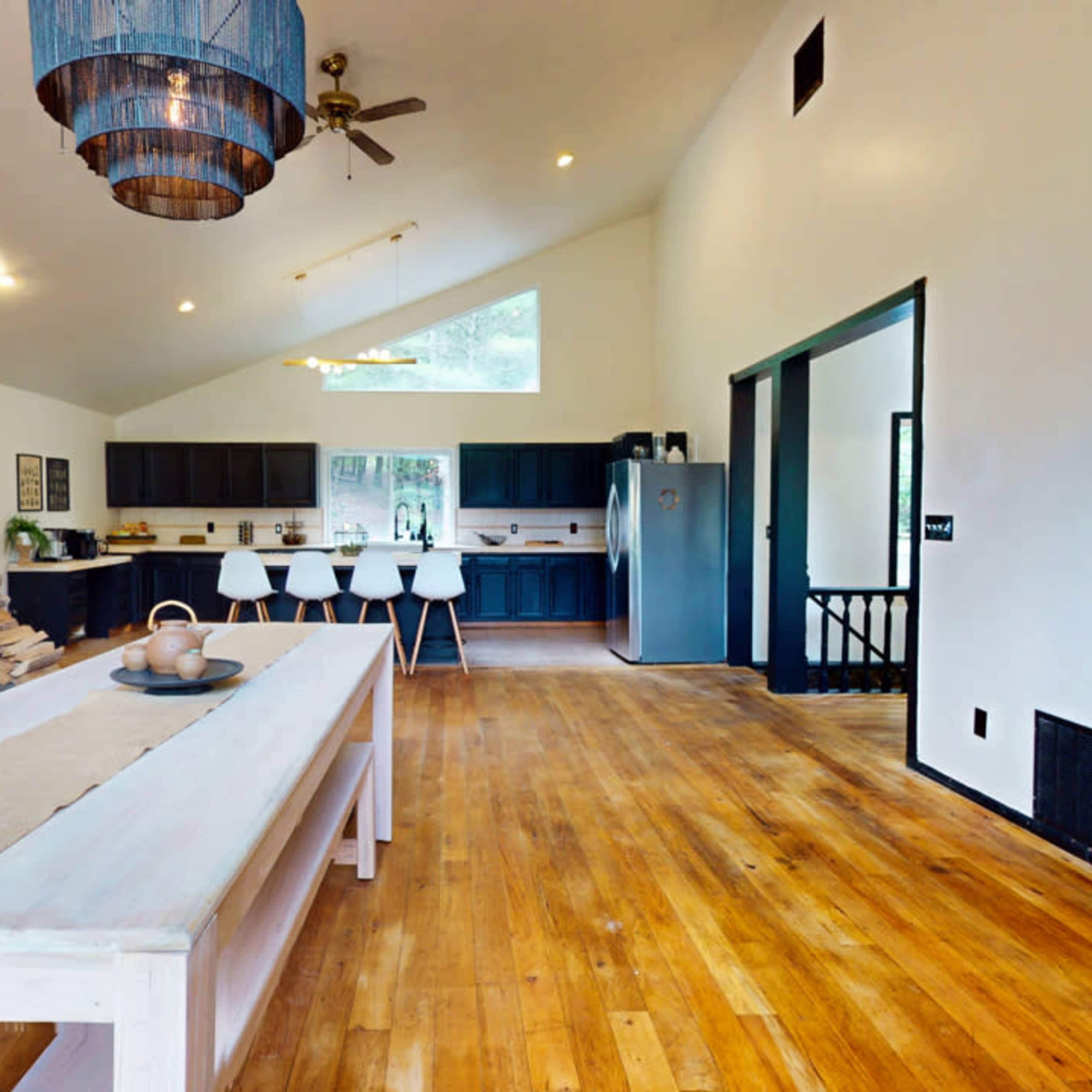 A modern kitchen with a wooden dining table, a stone fireplace, and dark cabinetry complemented by white walls and natural wood flooring.