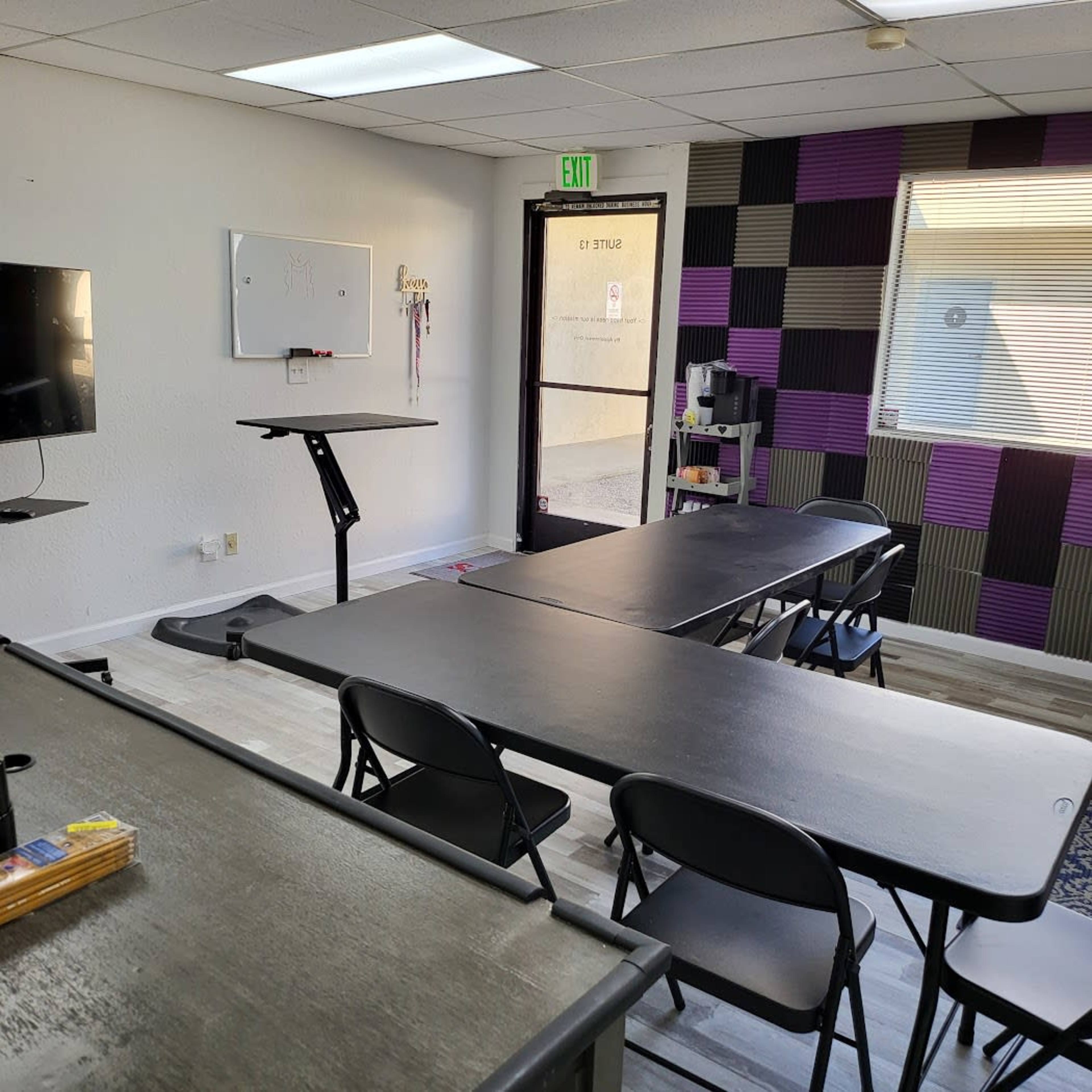 A meeting room with several black folding tables and chairs arranged, a wall-mounted TV, and a whiteboard.