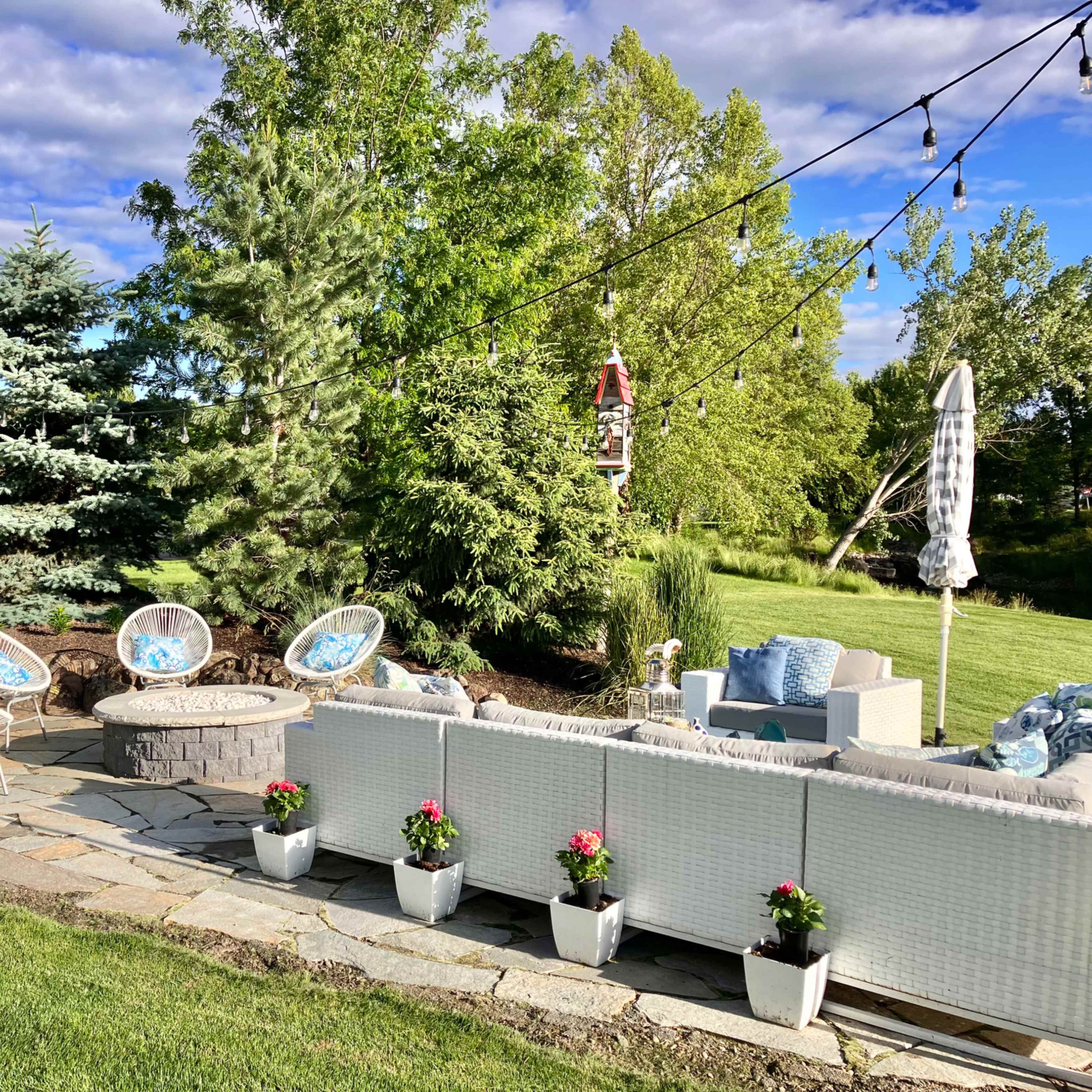 A landscaped outdoor patio area featuring a circular stone fire pit surrounded by four blue-accented chairs and a white sectional sofa, with potted flowers arranged along the patio stones.