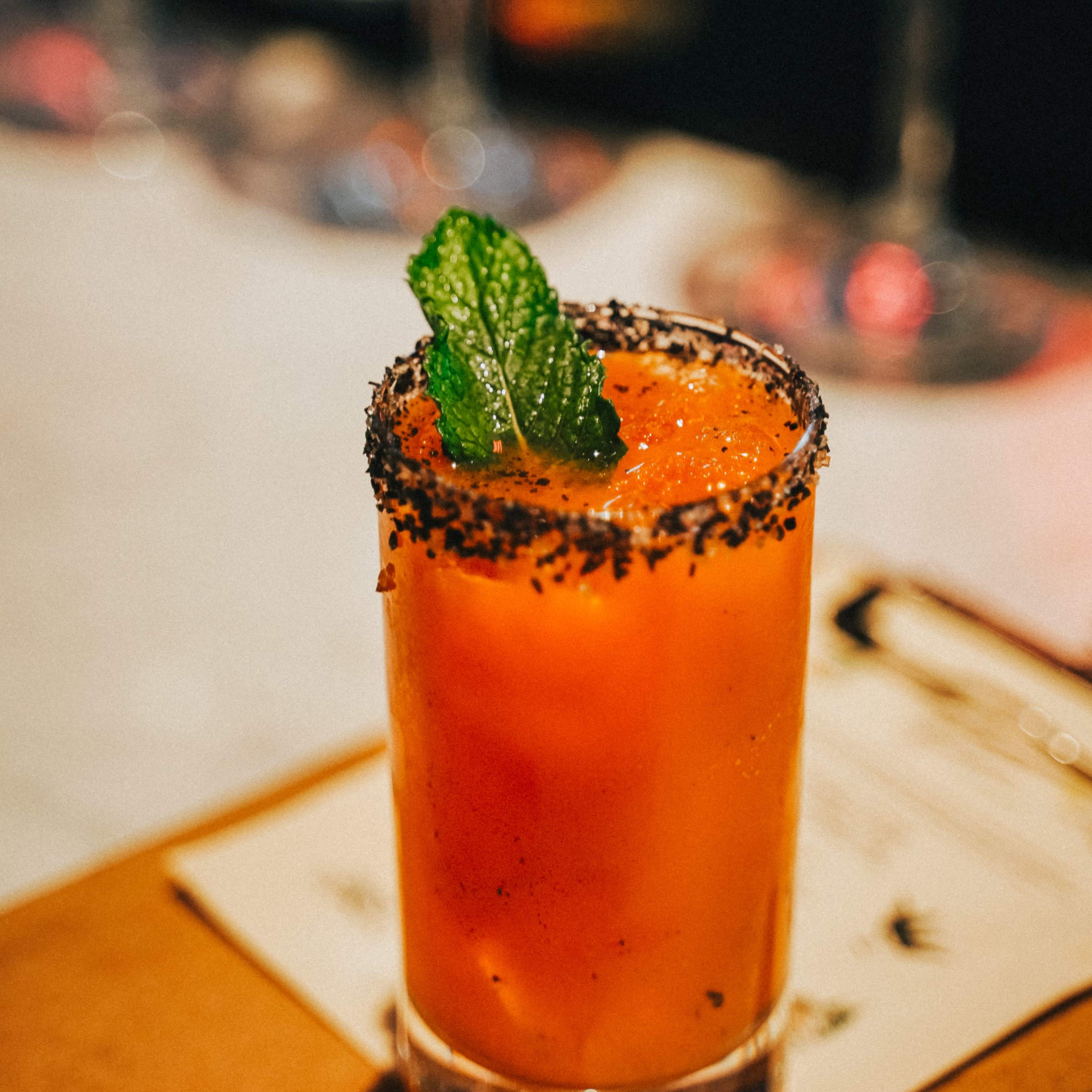 A glass of vibrant orange juice garnished with a mint leaf and a black salt rim sits on a wooden surface.