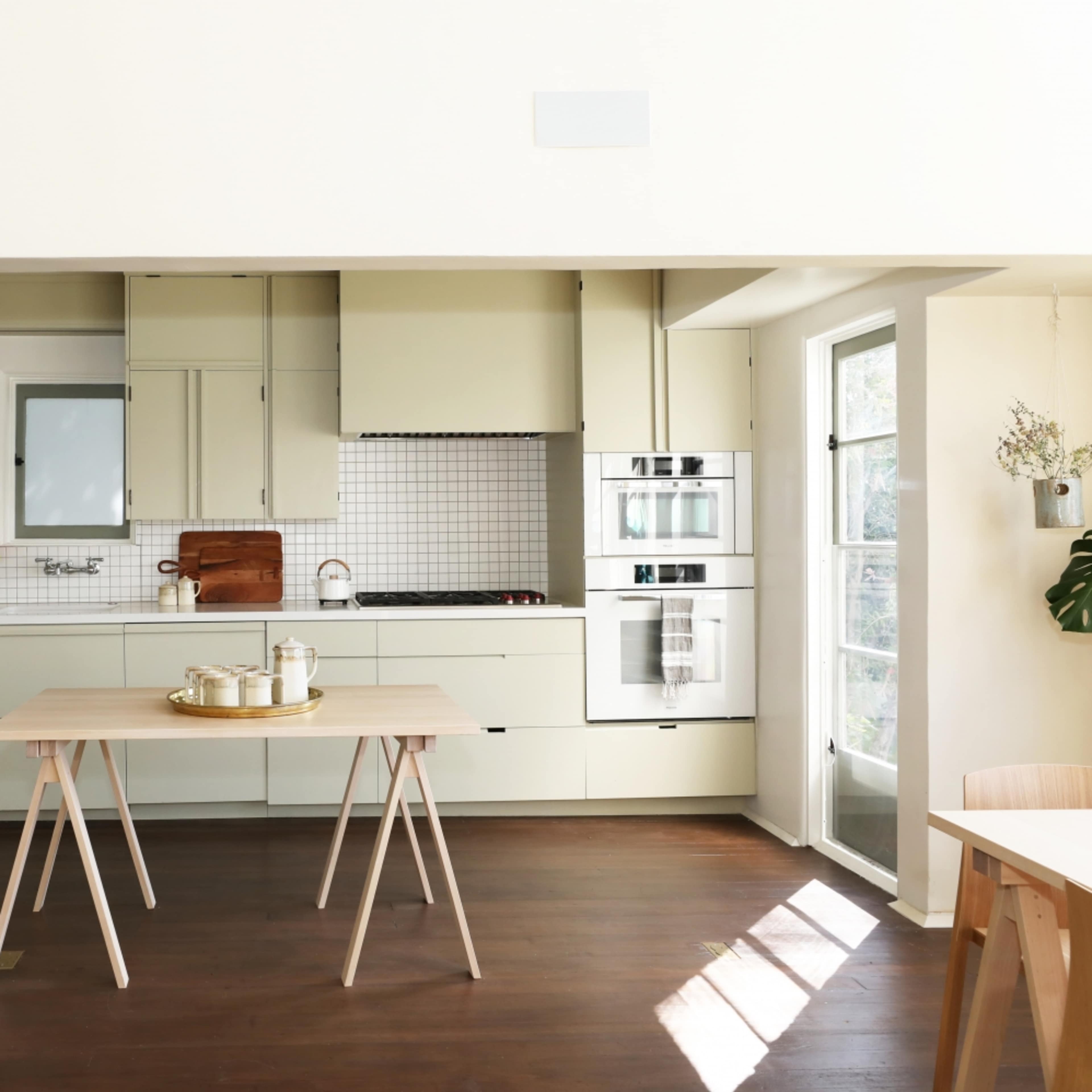 A modern kitchen with light green cabinets, a stainless steel refrigerator, and a wooden dining table set in the center.