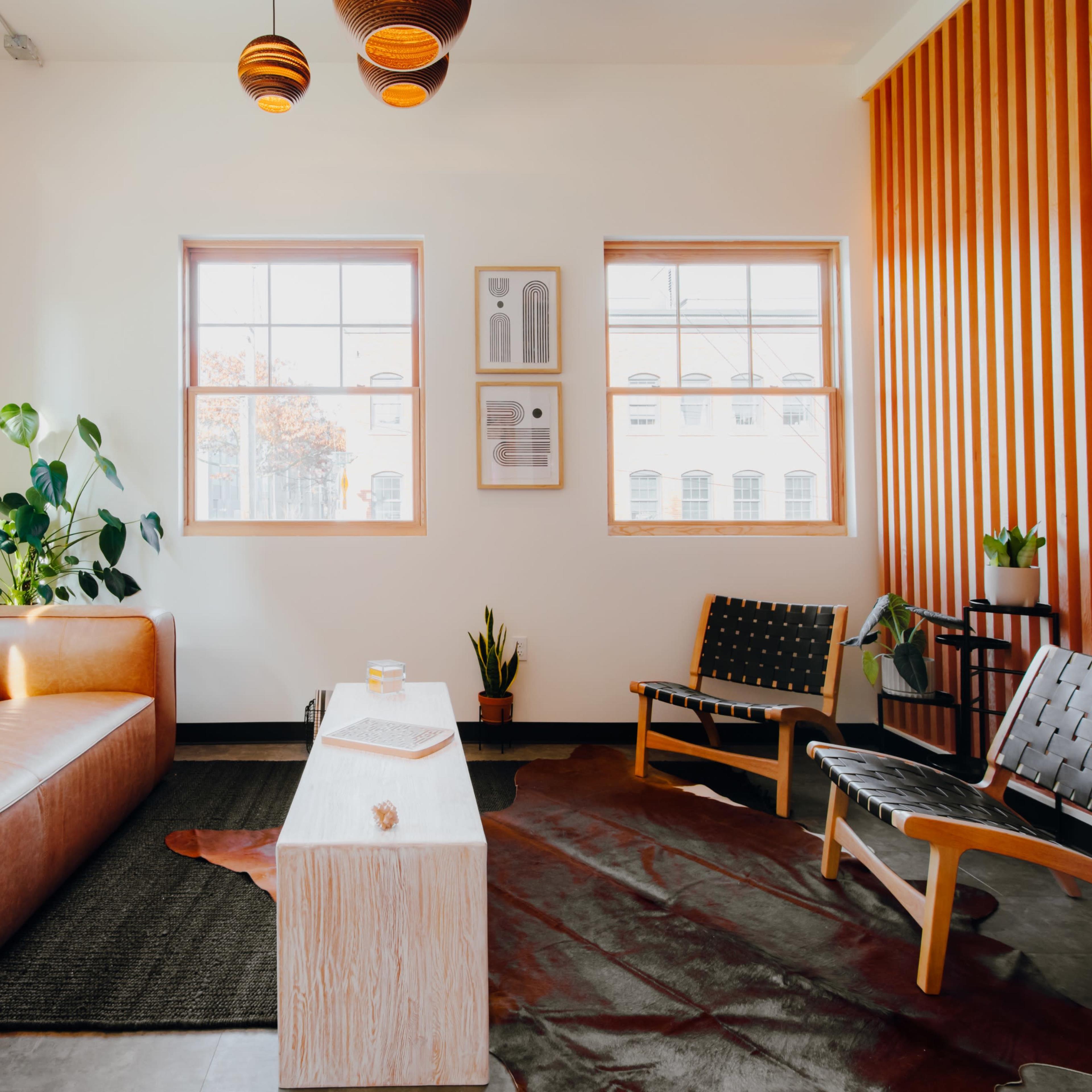 A modern living room featuring a brown leather sofa, a wooden coffee table, and various plants near large windows.