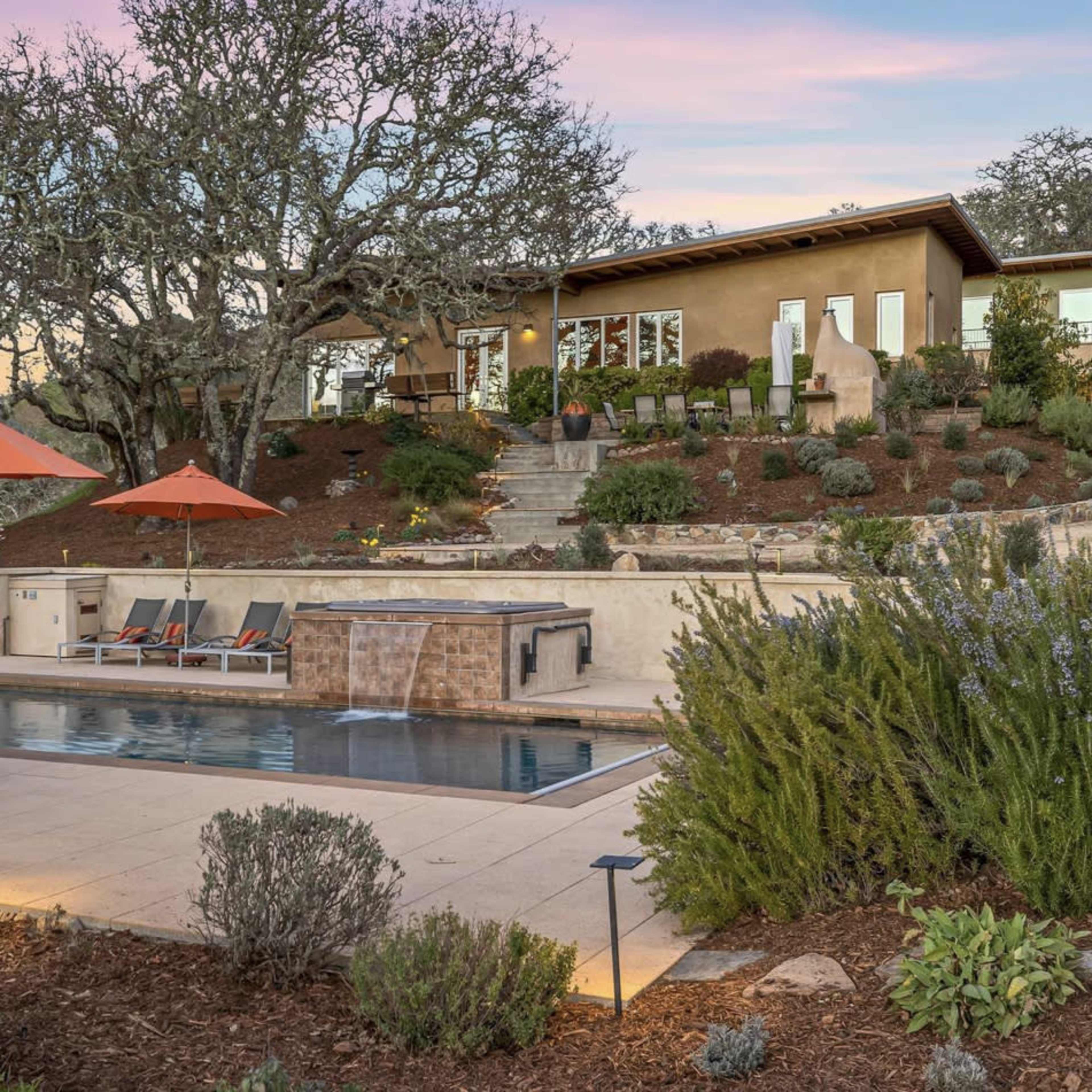 A modern outdoor area with a swimming pool, lounge chairs, and orange umbrellas, surrounded by landscaped gardens and a house in the background.