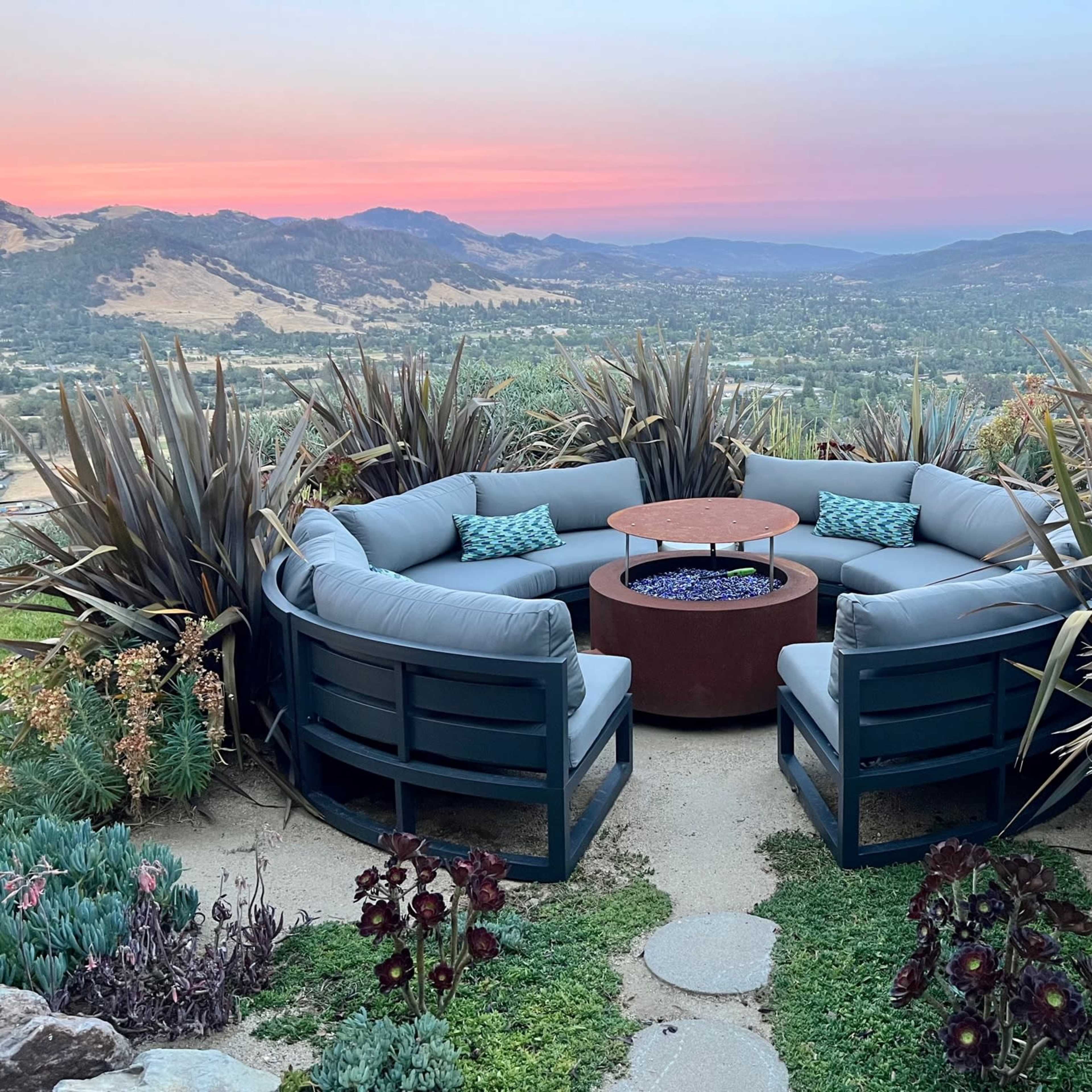 A circular seating area with blue cushions surrounds a central fire pit, set against a backdrop of mountains and valleys at sunset.