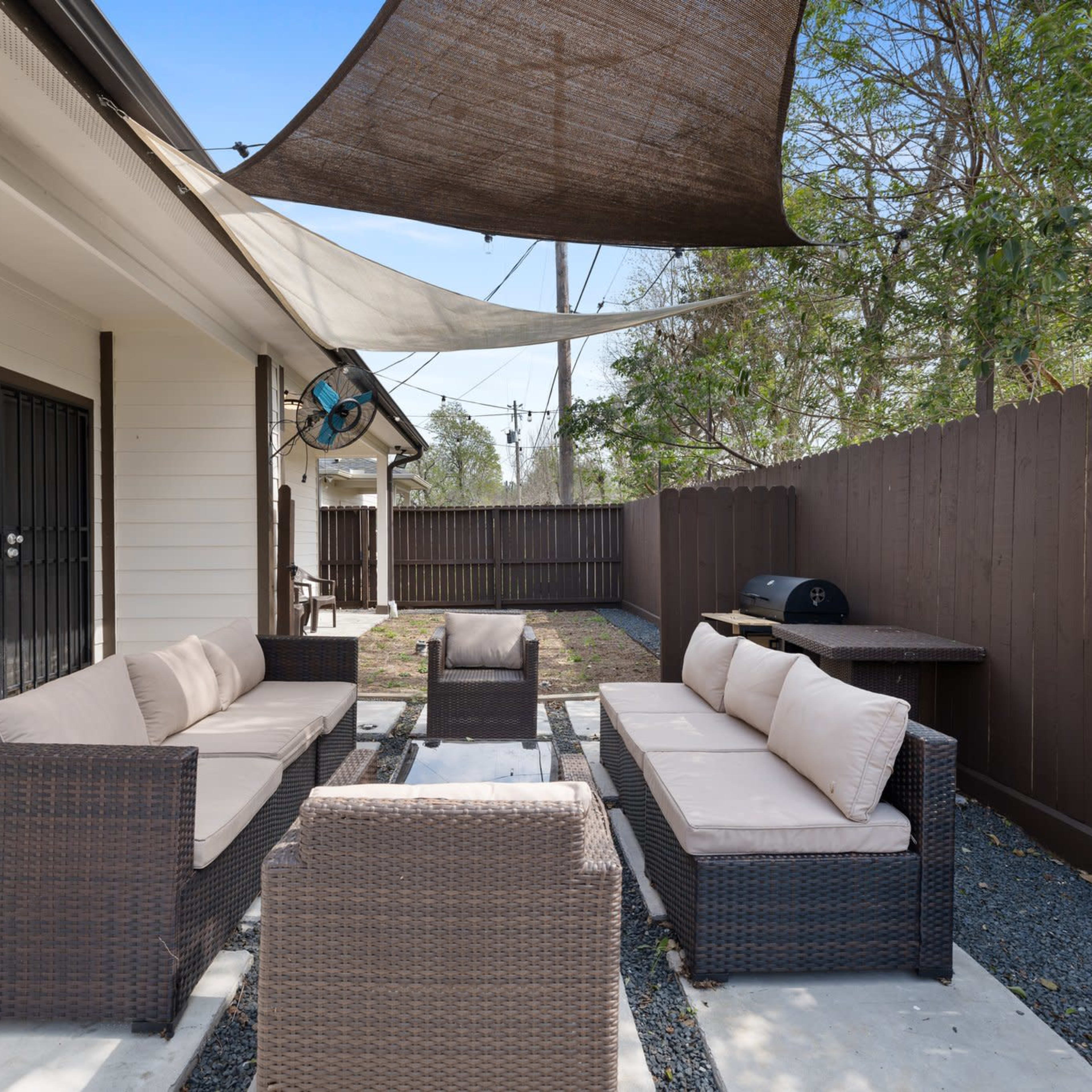 An outdoor seating area with a dark wicker sofa set, a glass-top table, a barbecue grill, and shaded by fabric canopies.