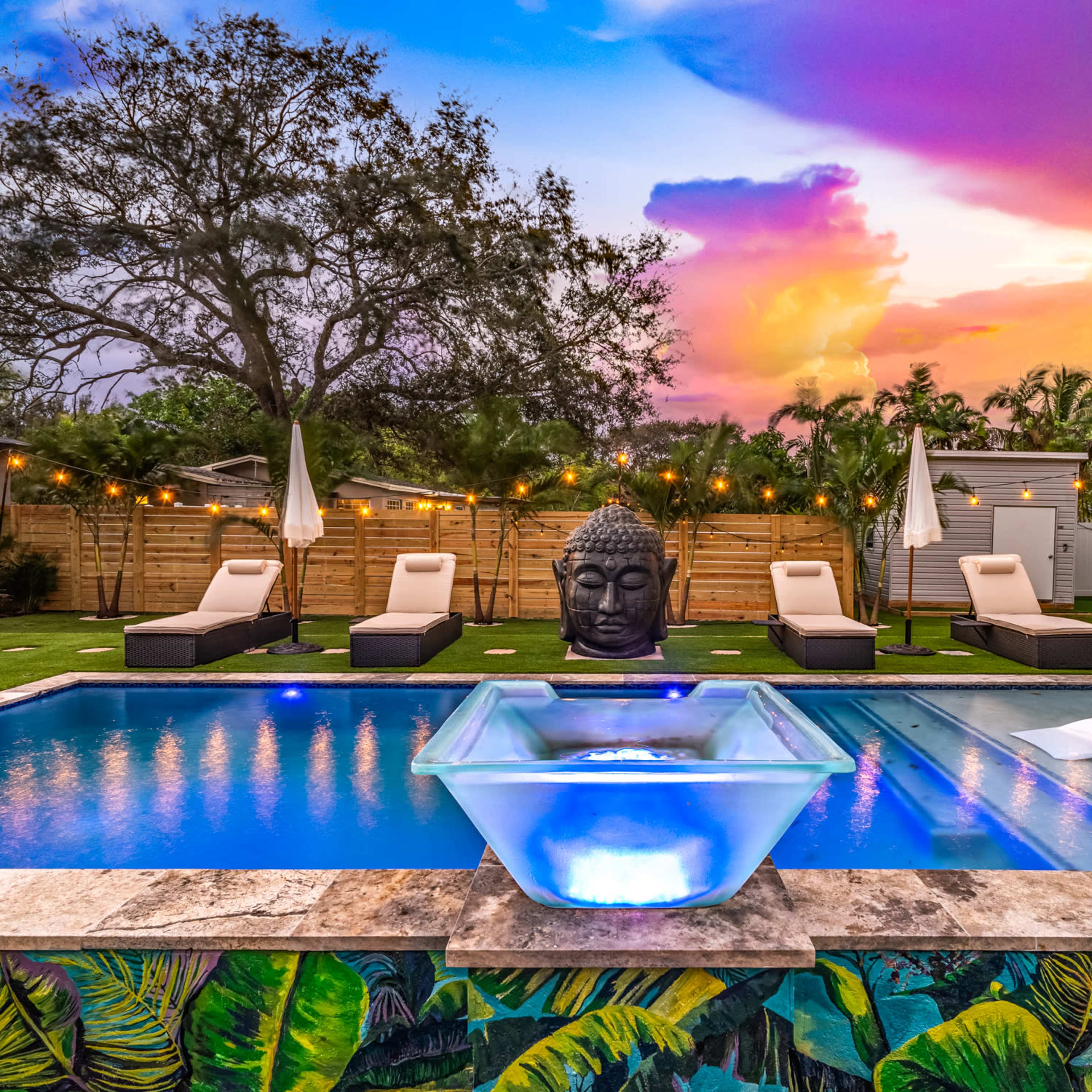A modern backyard features a swimming pool with a fountain, lounge chairs, a large Buddha statue, and vibrant sunset skies above.