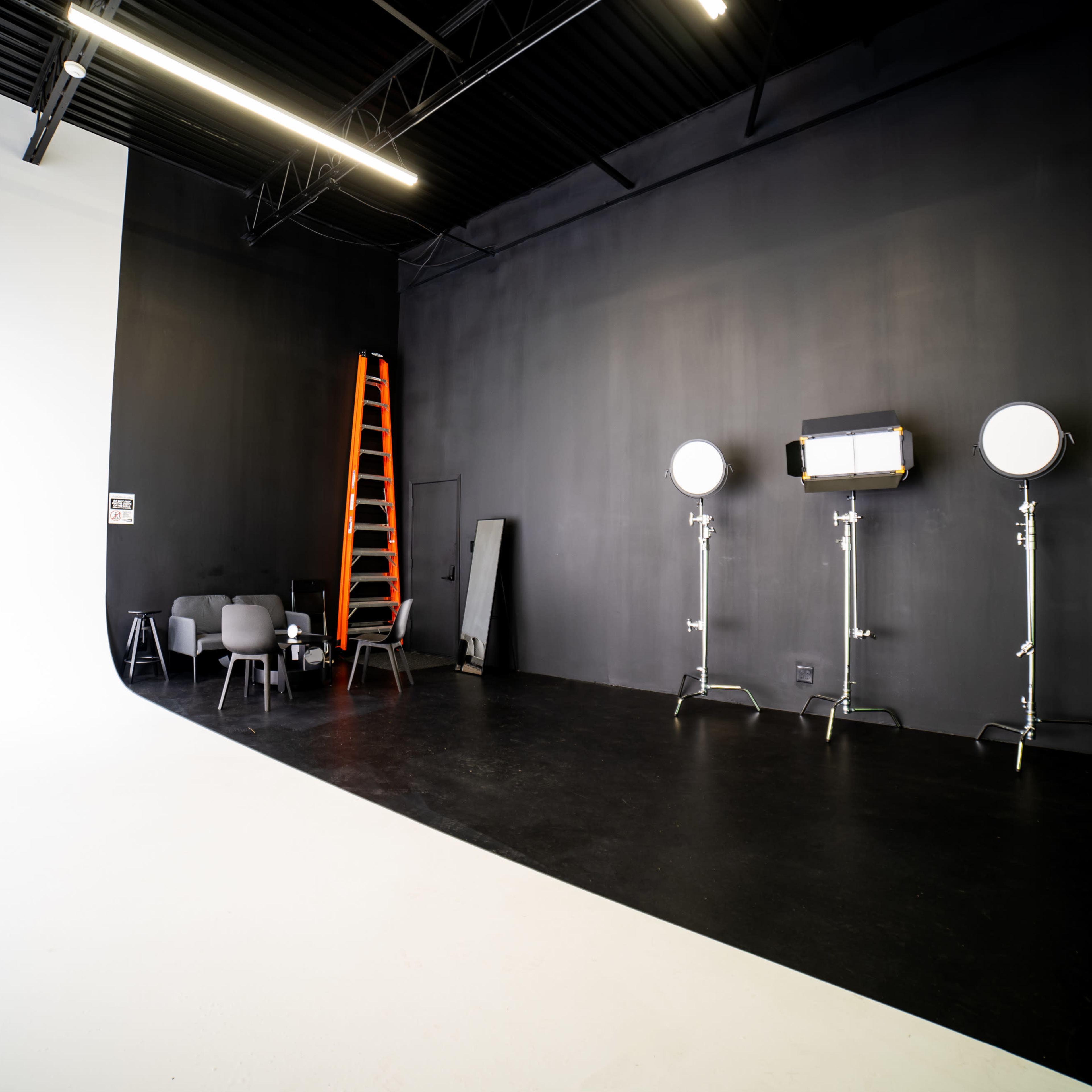A photography studio features a white backdrop on one side, black walls, a ladder, lighting equipment, and a few chairs.