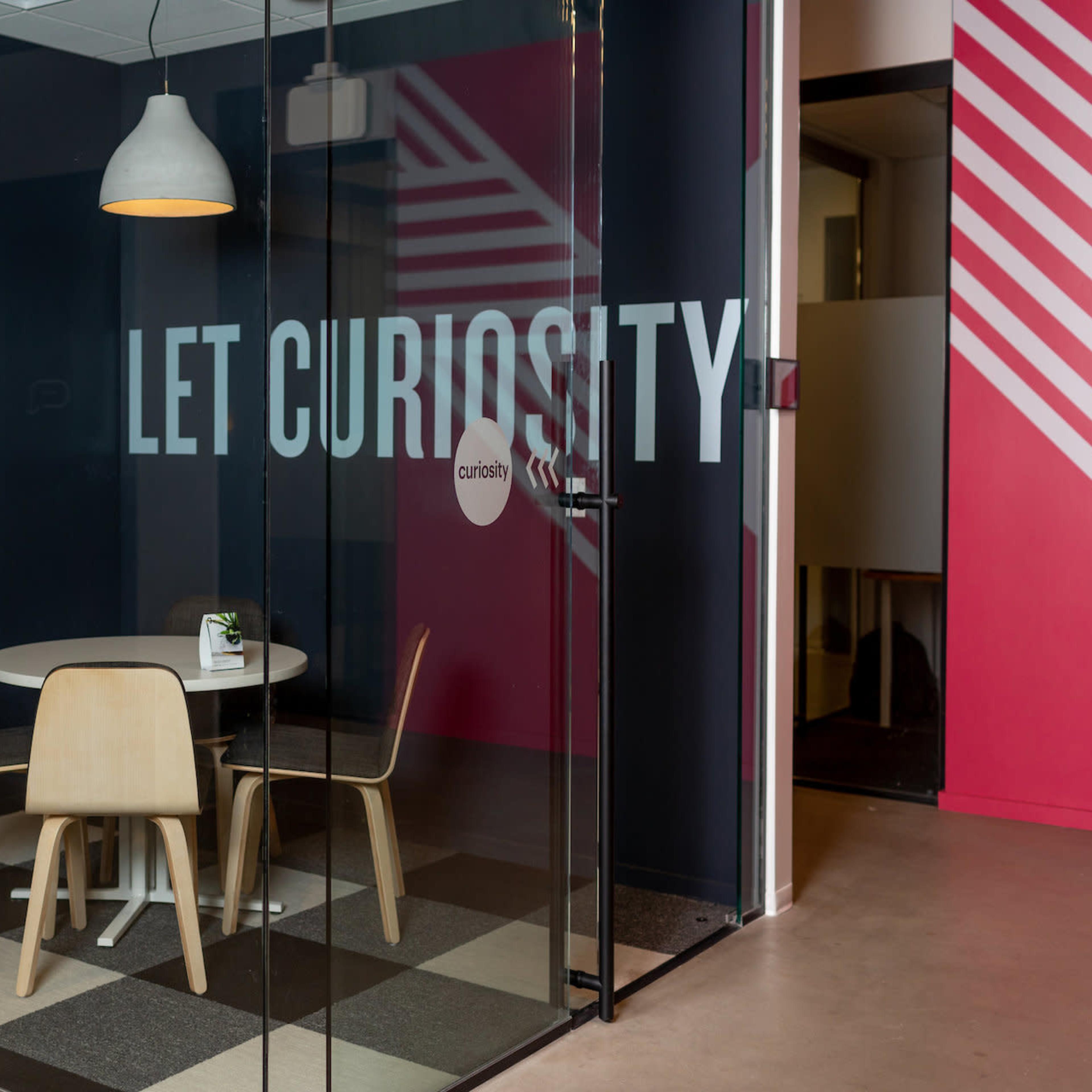 A modern glass conference room with a round table and wooden chairs, featuring a wall with the phrase "LET CURIOSITY" prominently displayed.
