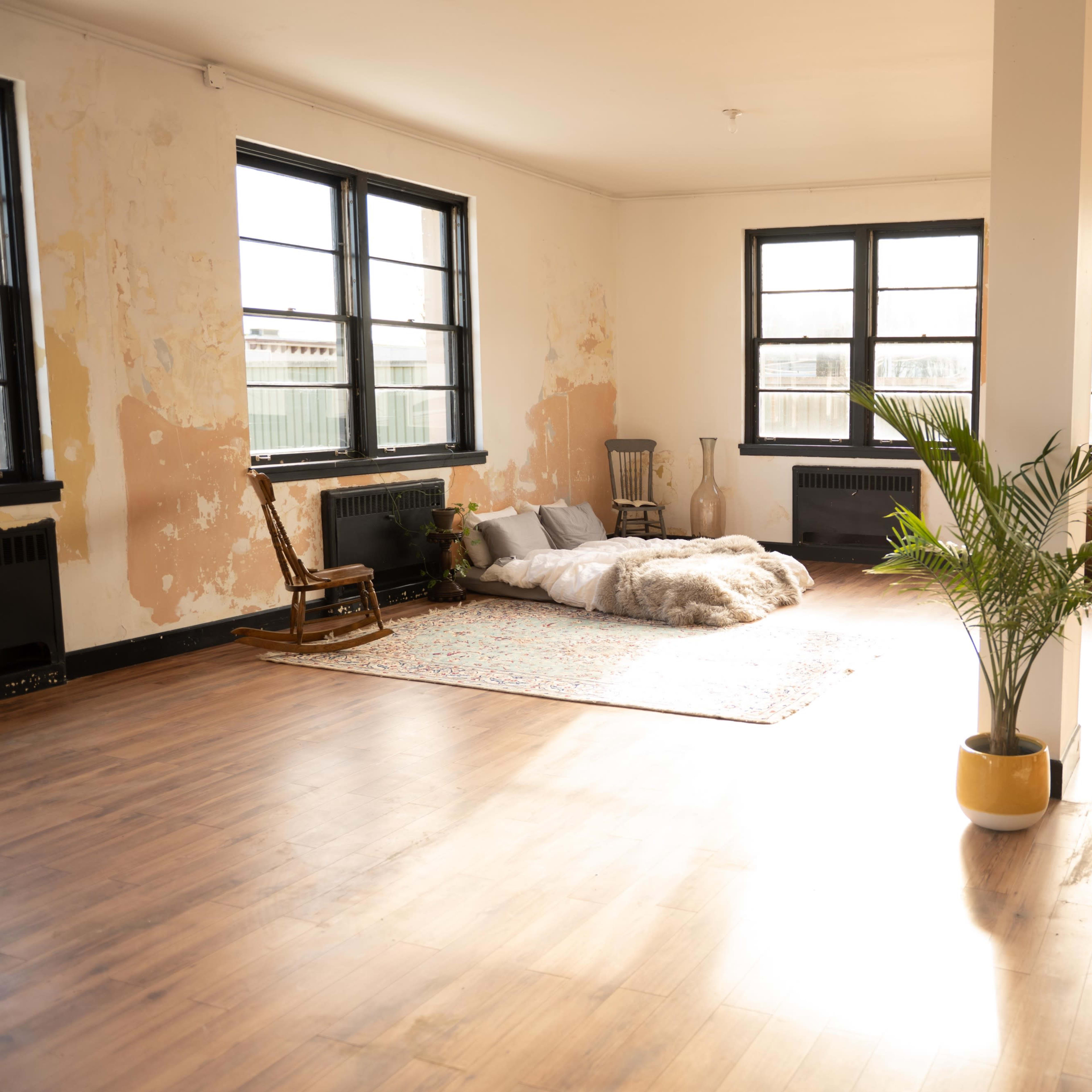 A spacious, partially furnished room with wooden floors, large windows, a soft area for sitting, and a potted plant.