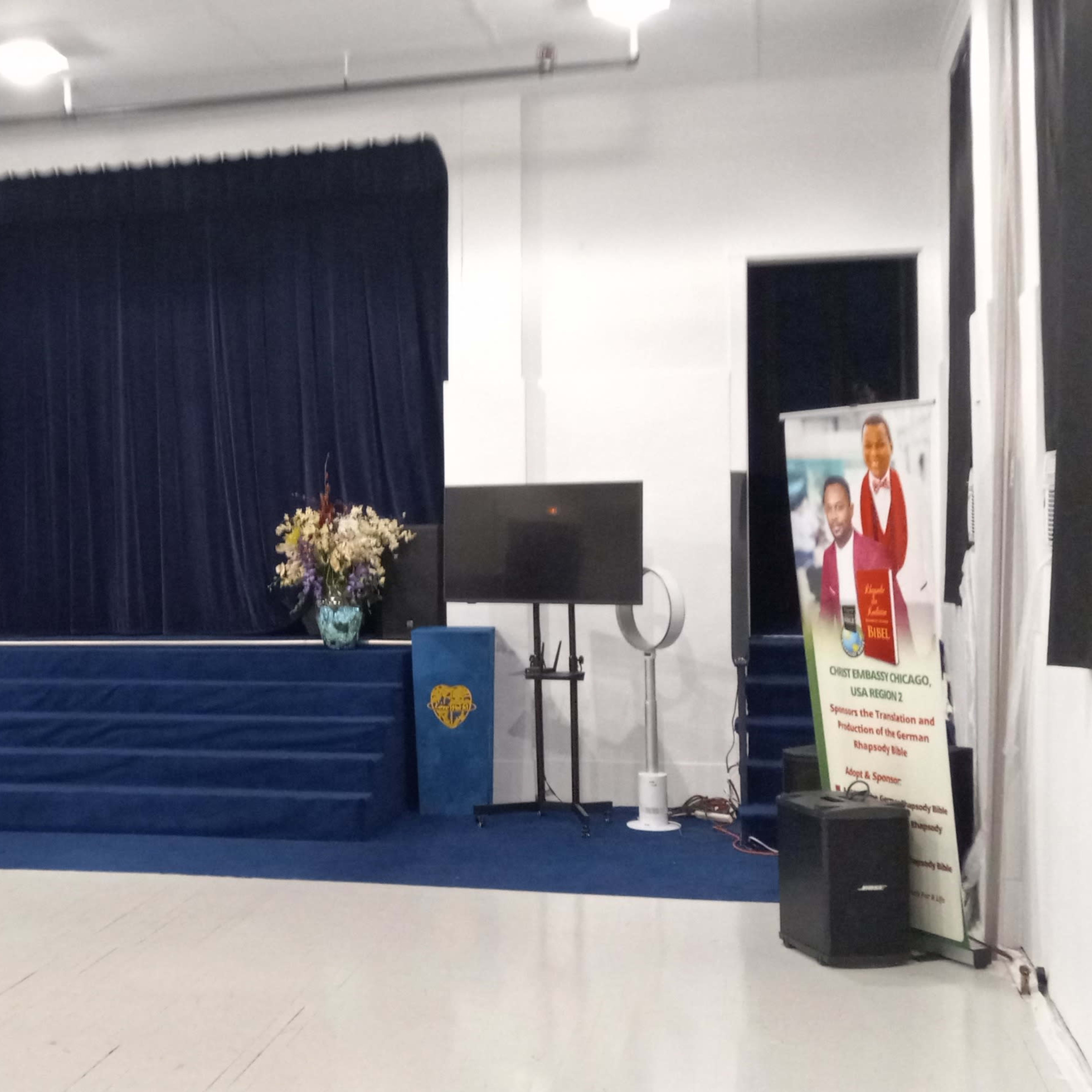 A stage with blue curtains, flanked by monitors and decorative flower arrangements, set in a well-lit hall with a blue carpeted floor.
