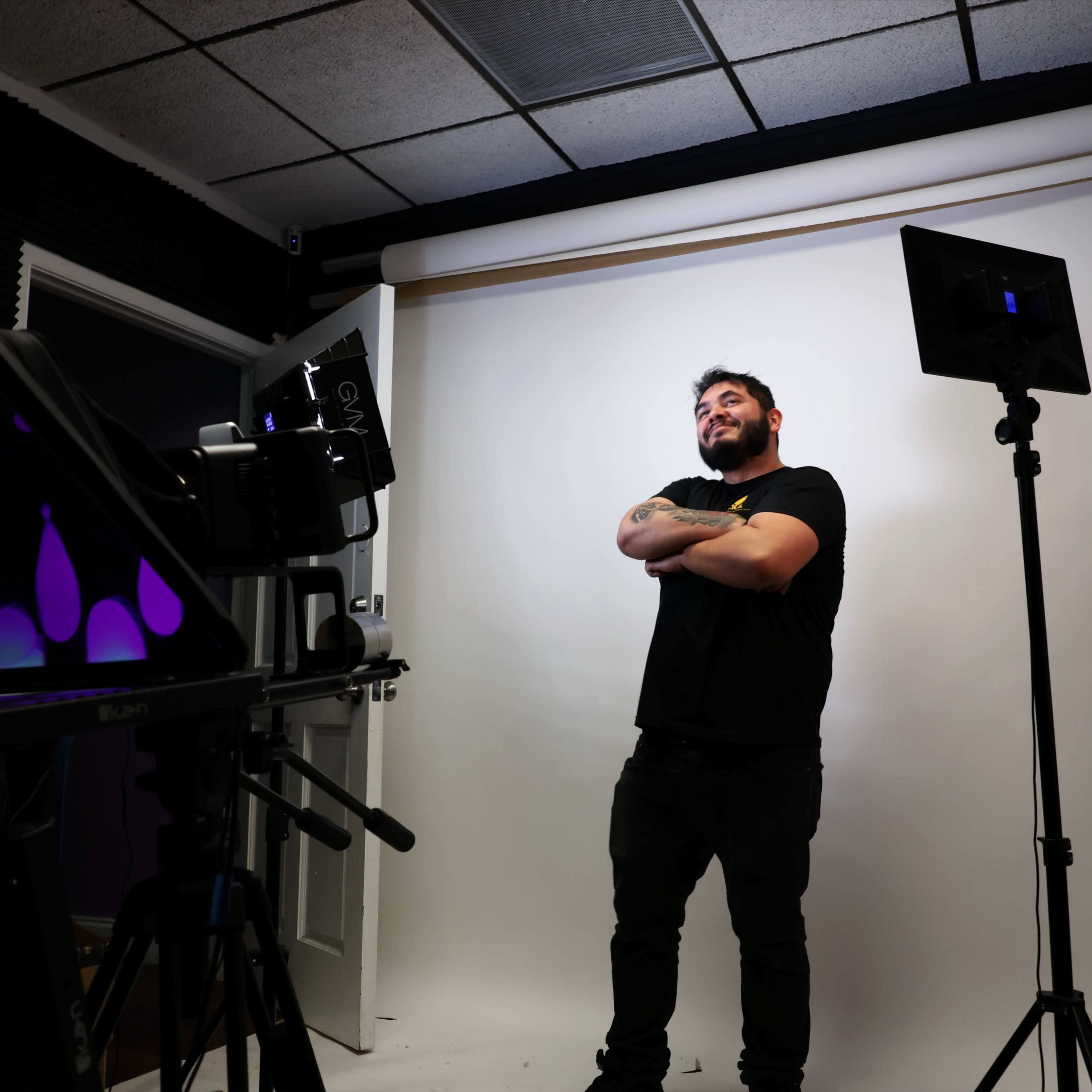 A man stands confidently with his arms crossed in front of a plain white backdrop, surrounded by film and lighting equipment.