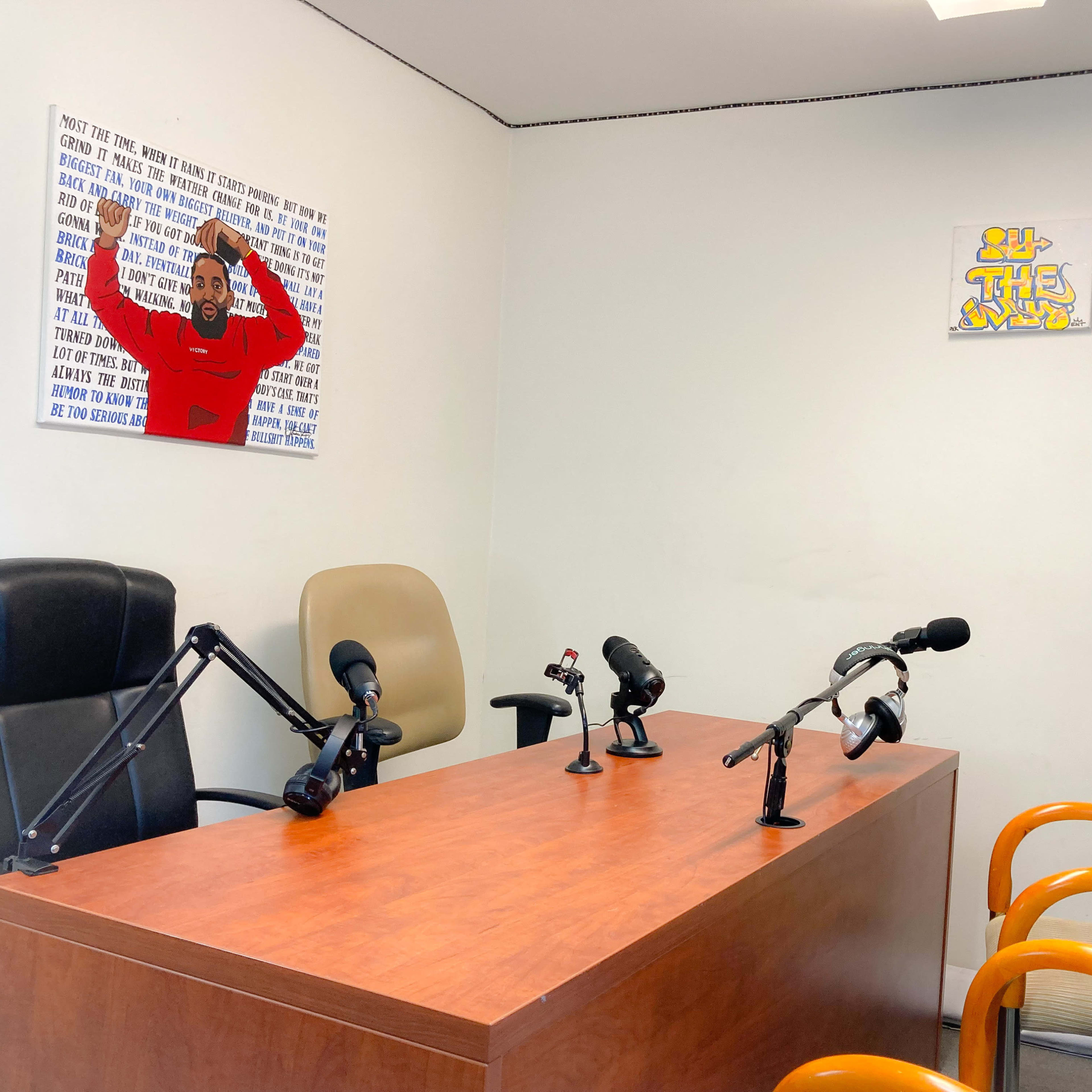 A small recording studio features a wooden table with several microphones and chairs against a white wall adorned with two pieces of art.