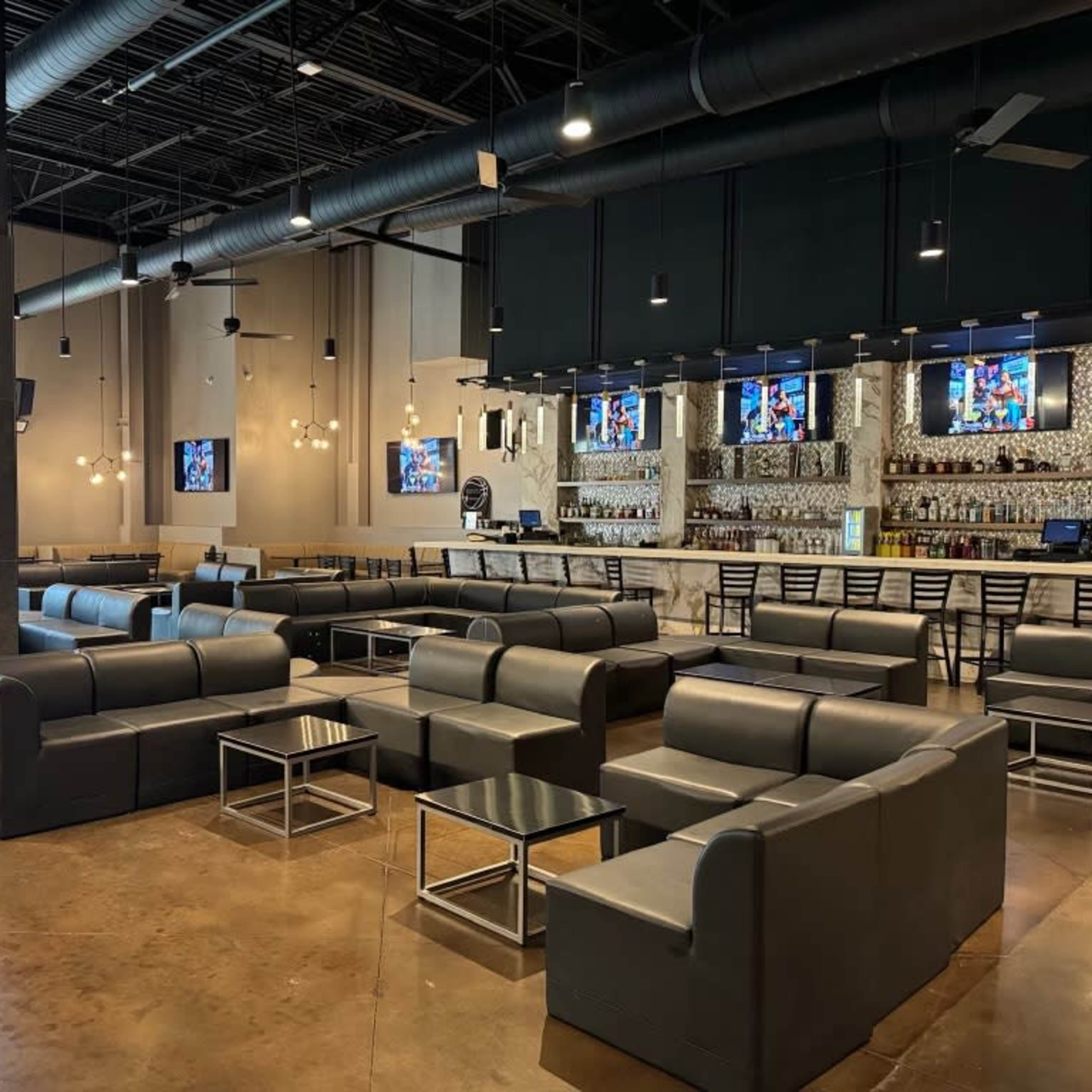 A spacious, modern lounge area with gray sofas and a bar counter featuring multiple flat-screen TVs in a well-lit interior.