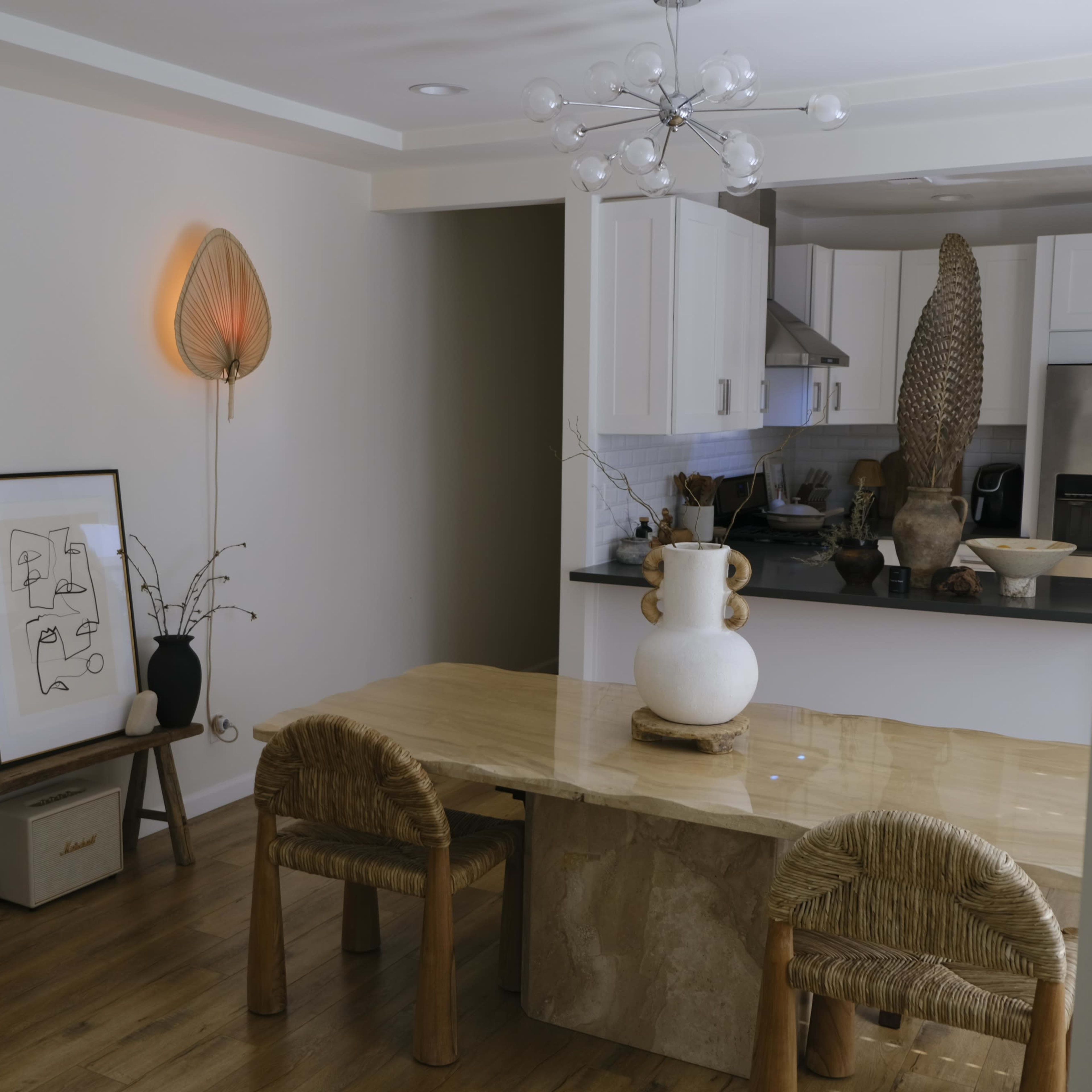 A modern kitchen and dining area featuring a wooden table, woven chairs, and decorative elements including wall sconces and a large vase.