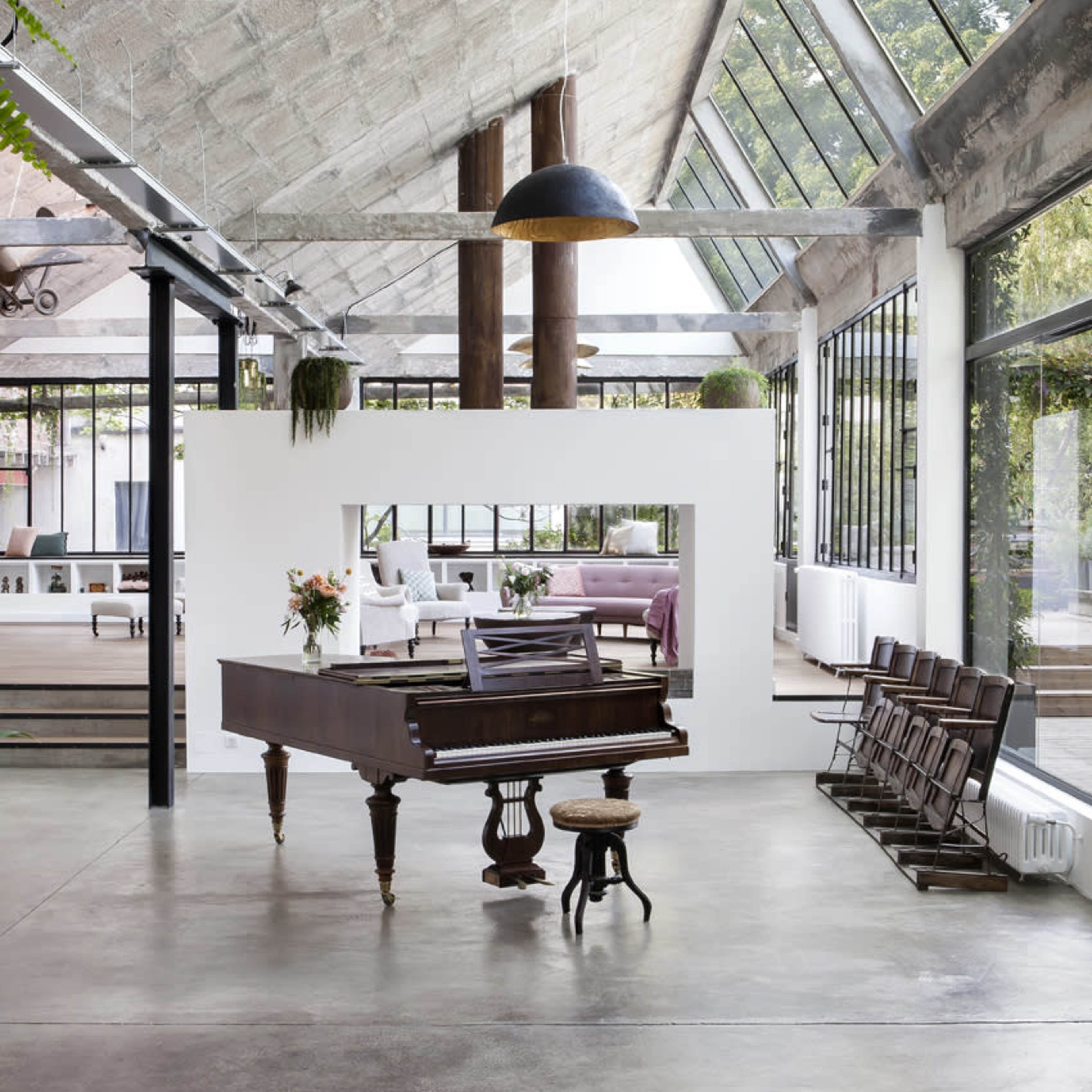A grand piano is positioned in the center of a spacious room with high ceilings, large windows, and industrial decor elements.