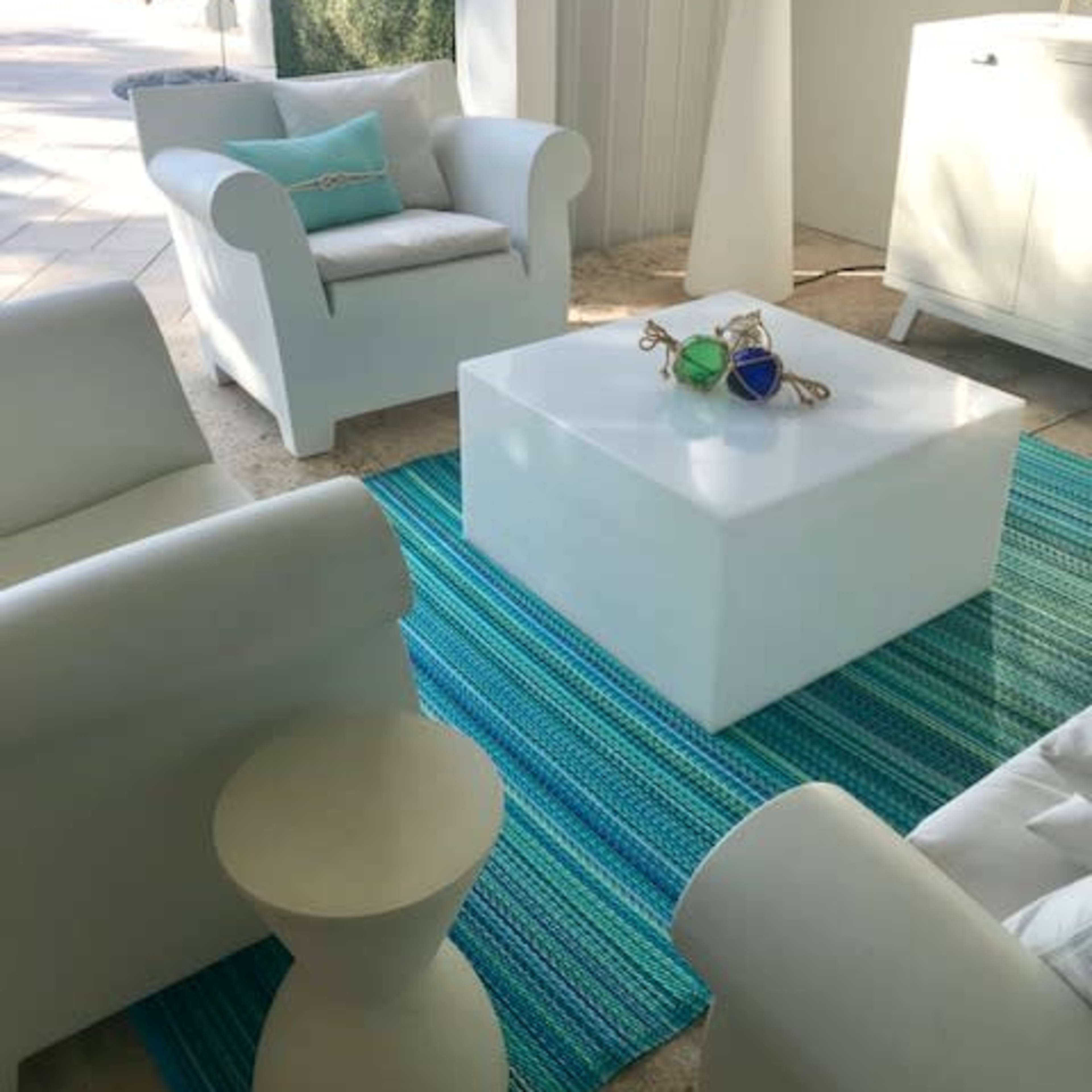 A modern lounge area features white furniture, including two chairs and a low table, on a blue and green striped rug, with a view of the ocean in the background.