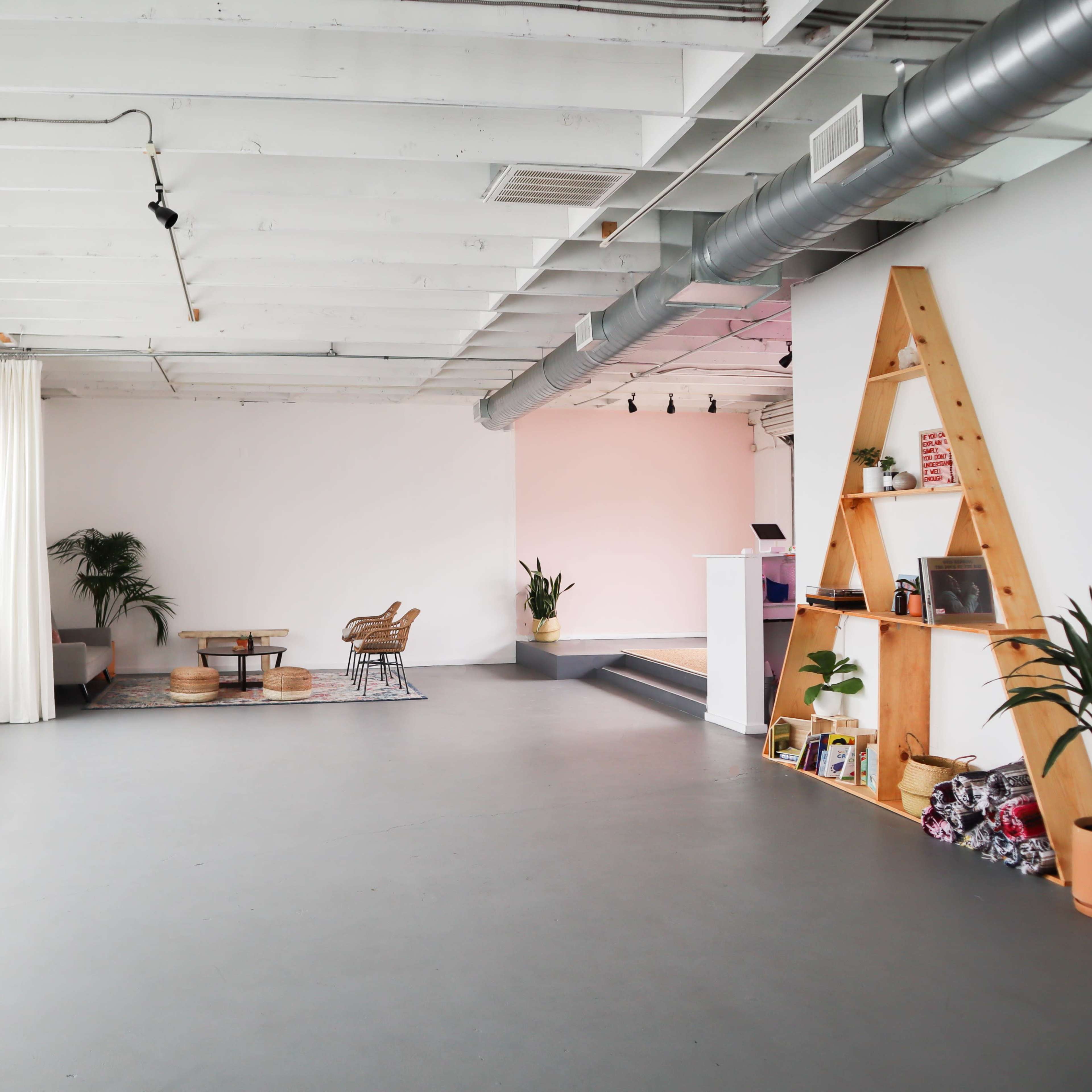 A spacious, minimalist room features large windows, plants, a seating area with chairs and a table, and a decorative shelving unit.