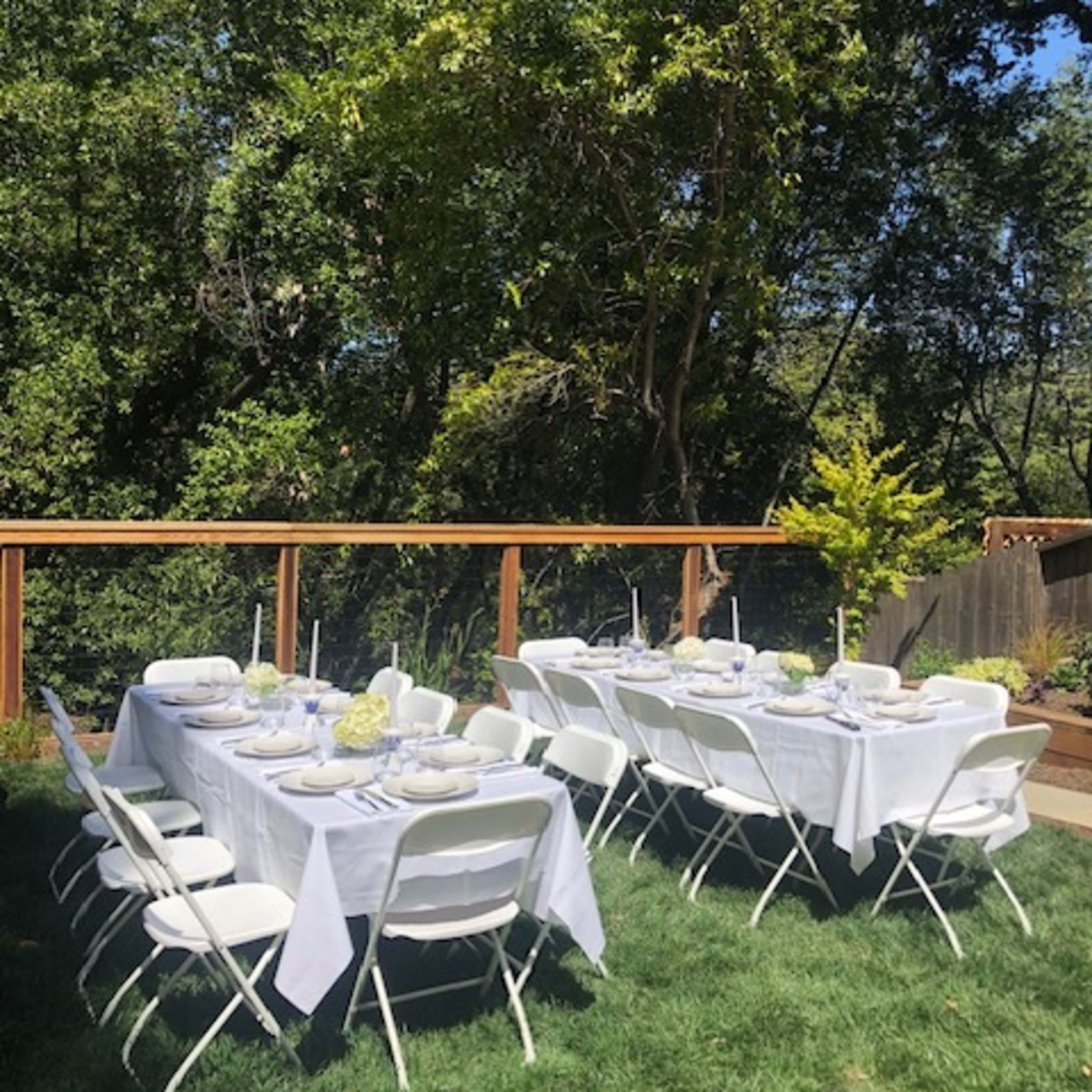 A long rectangular table is set with white tablecloths and dishes under a wooden deck surrounded by greenery.