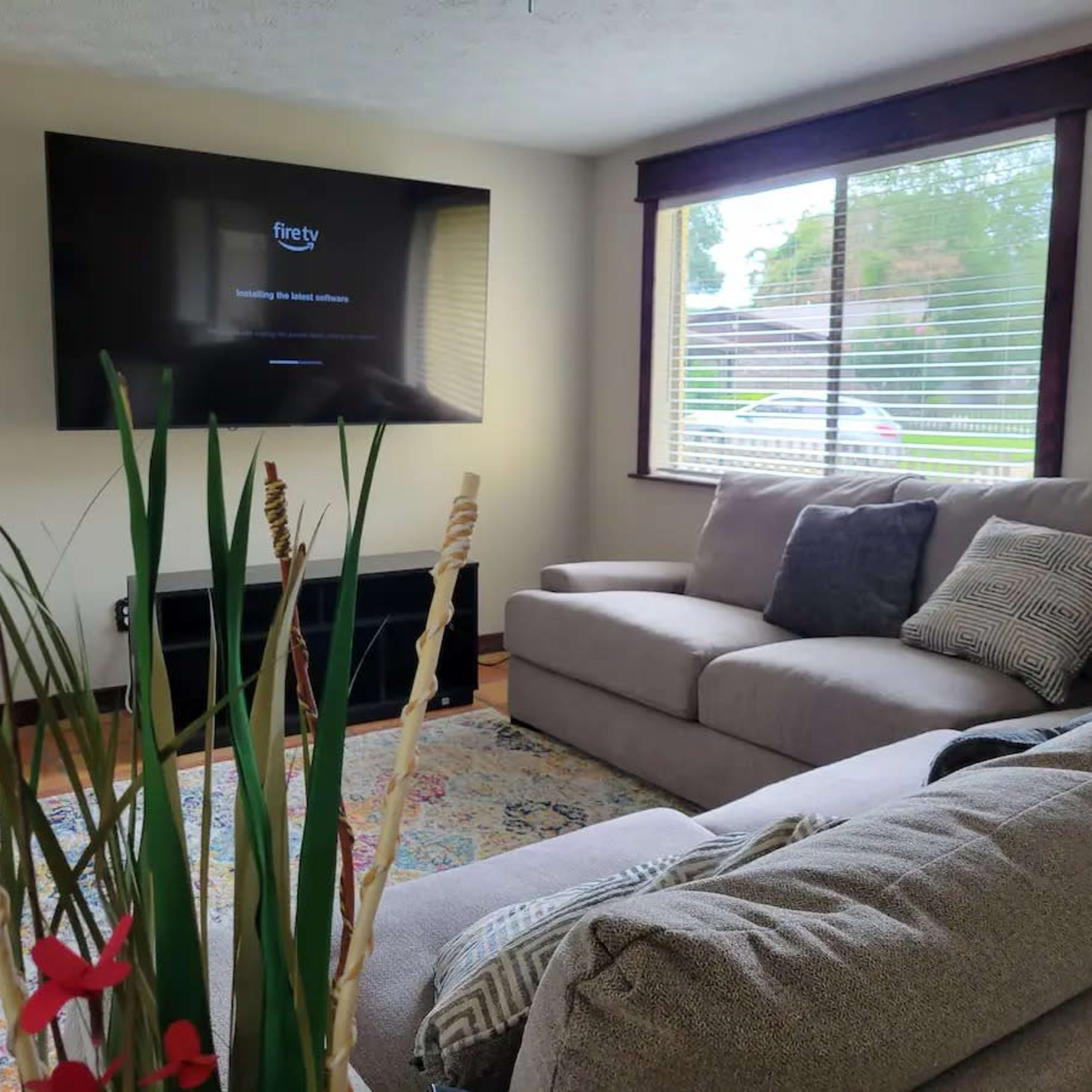 A living room with a sectional sofa, a television mounted on the wall, and a window with blinds letting in natural light.