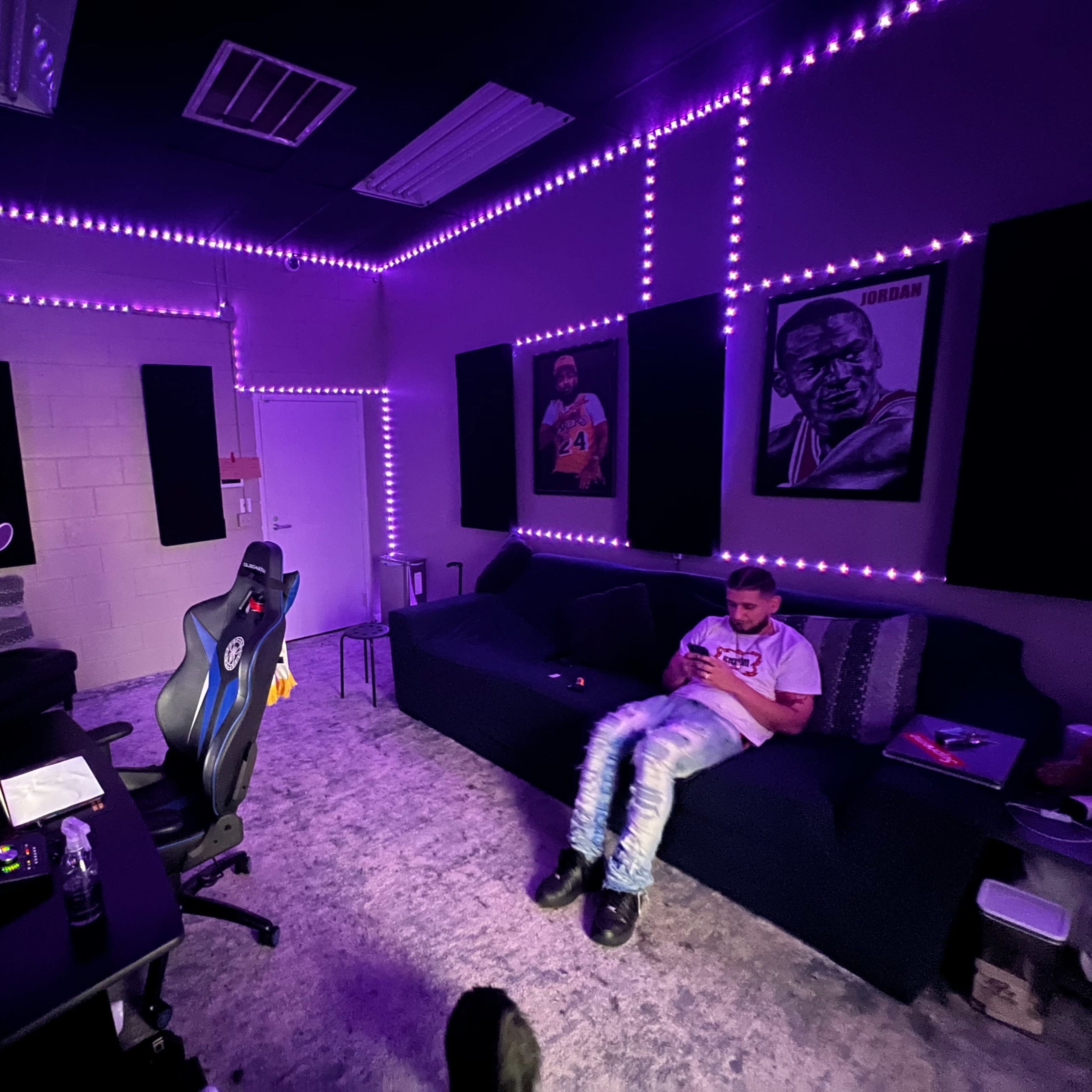 A dimly lit recording studio with purple LED lights, featuring a person sitting on a couch while looking at their phone.