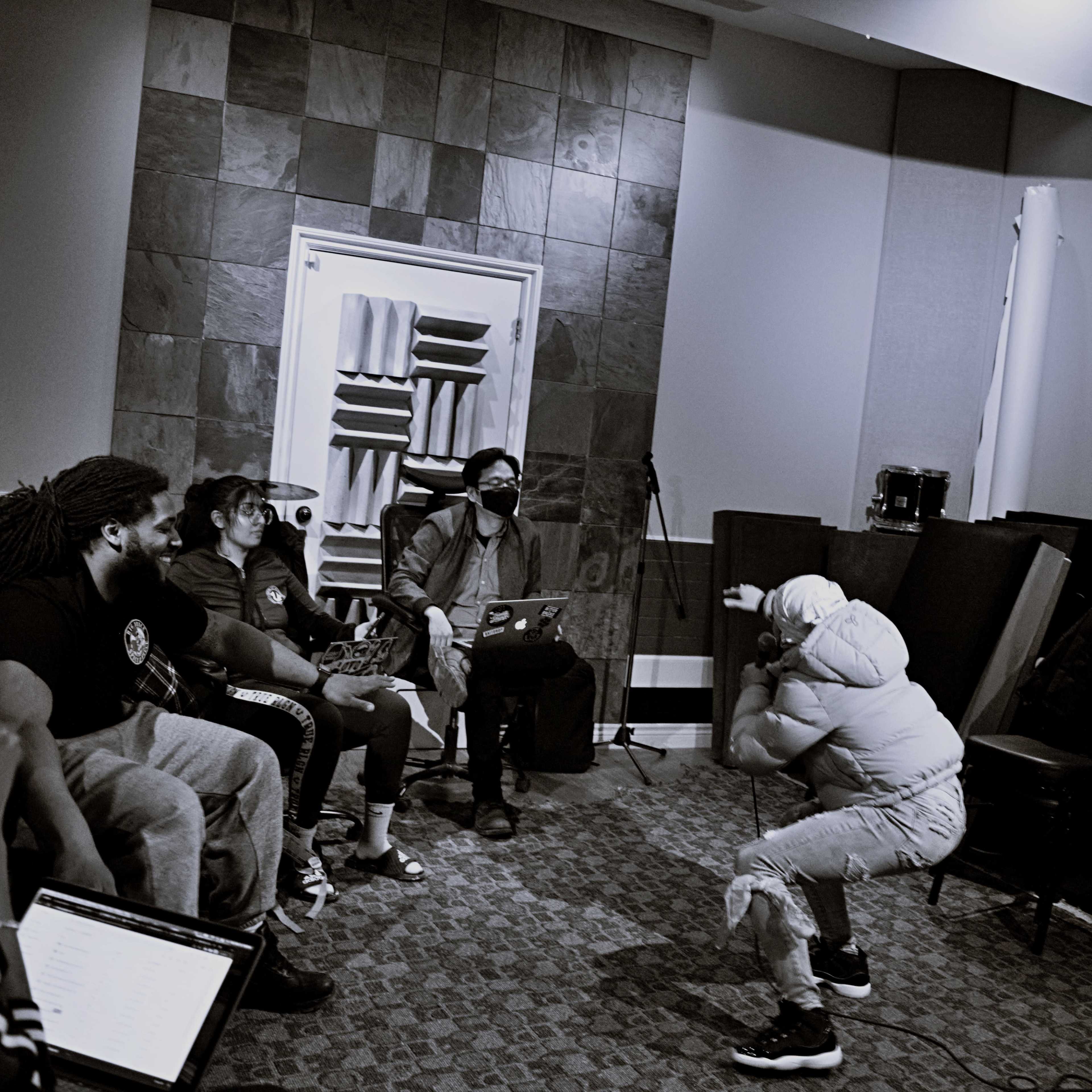 A group of six individuals watches a performer in a recording studio setting.