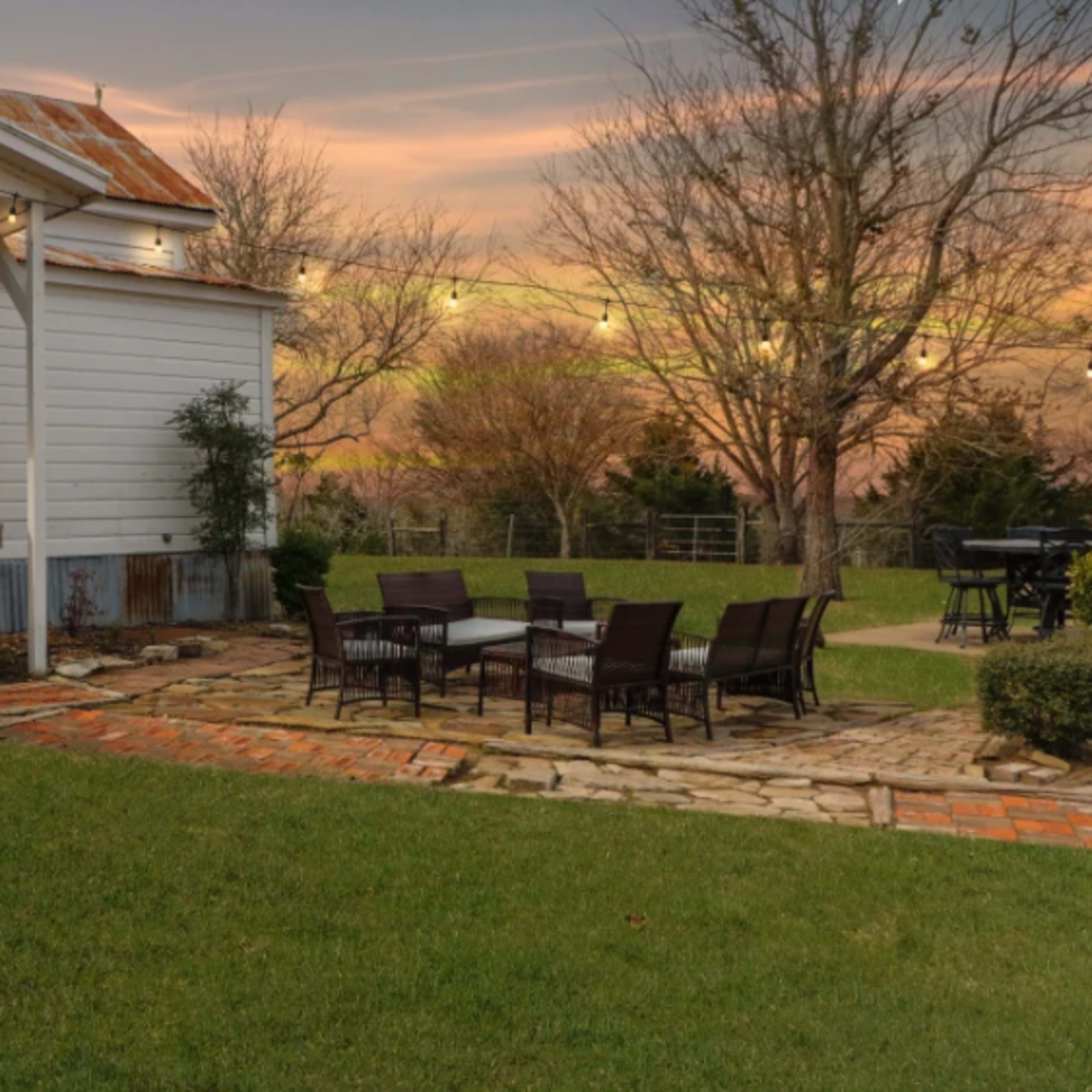 A backyard with a stone patio, chairs around a table, and a white house with a rustic metal roof at sunset.