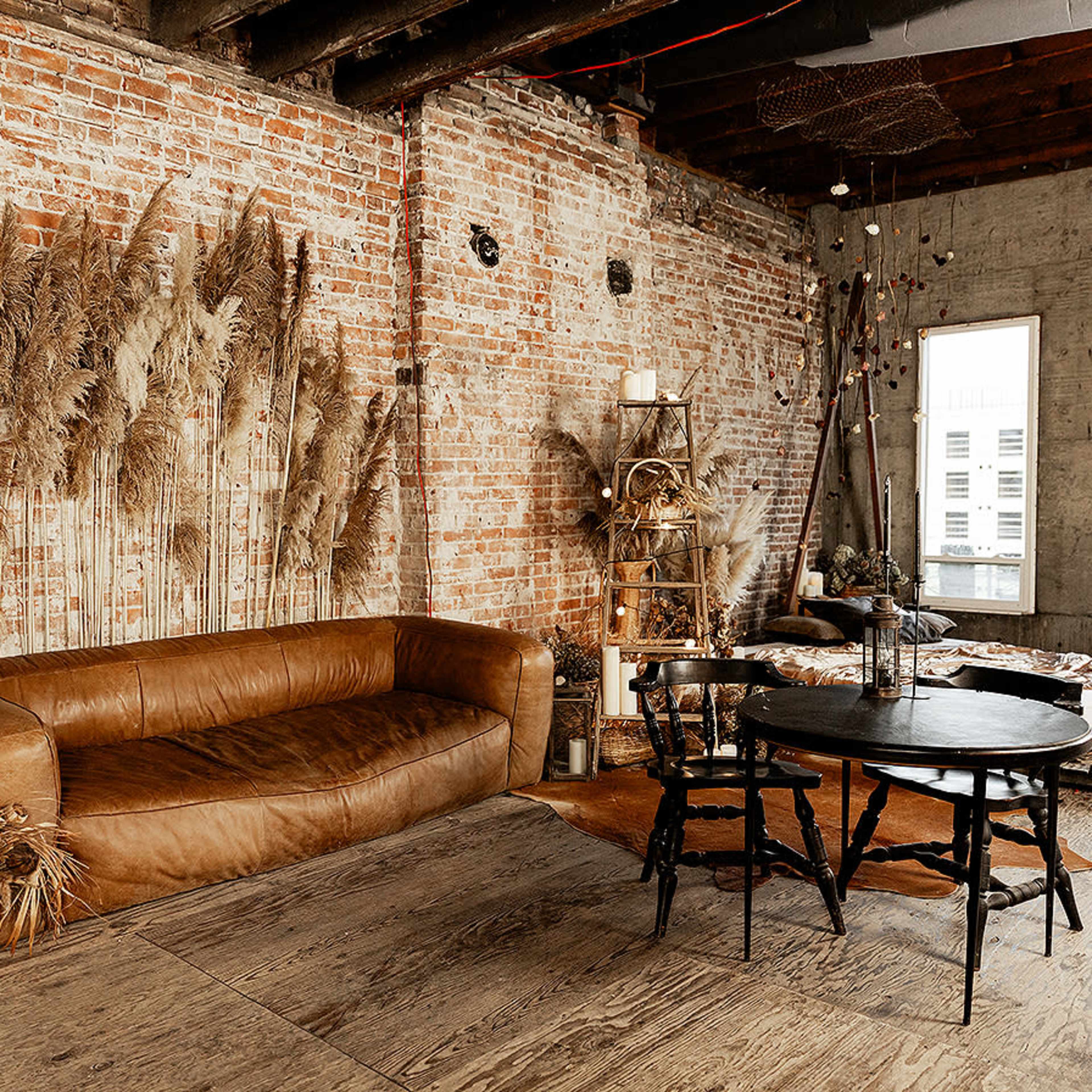 An industrial-style room with exposed brick walls, a brown leather sofa, a round black table, and large windows.