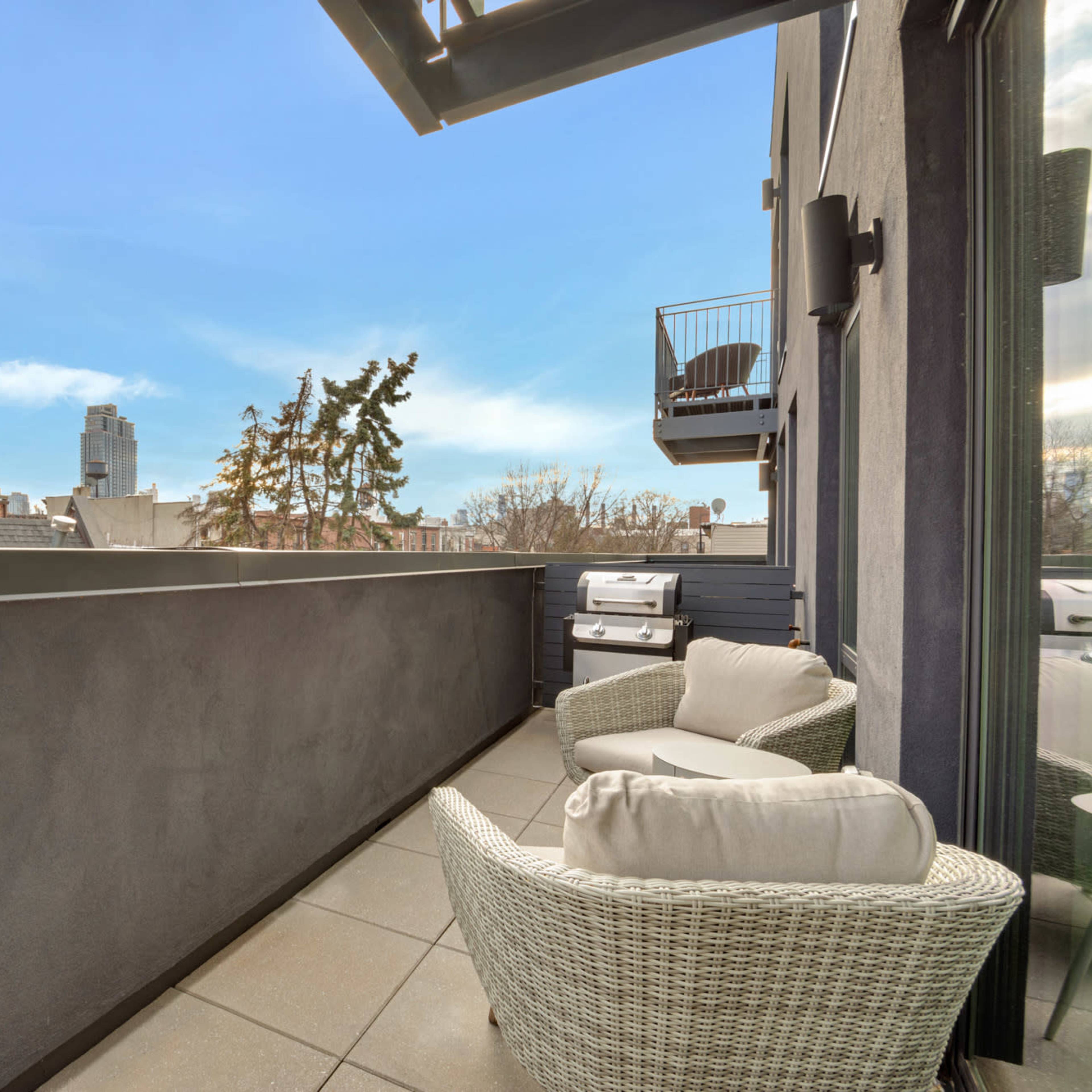 A modern balcony with two wicker chairs, overlooking rooftops and trees against a clear blue sky.