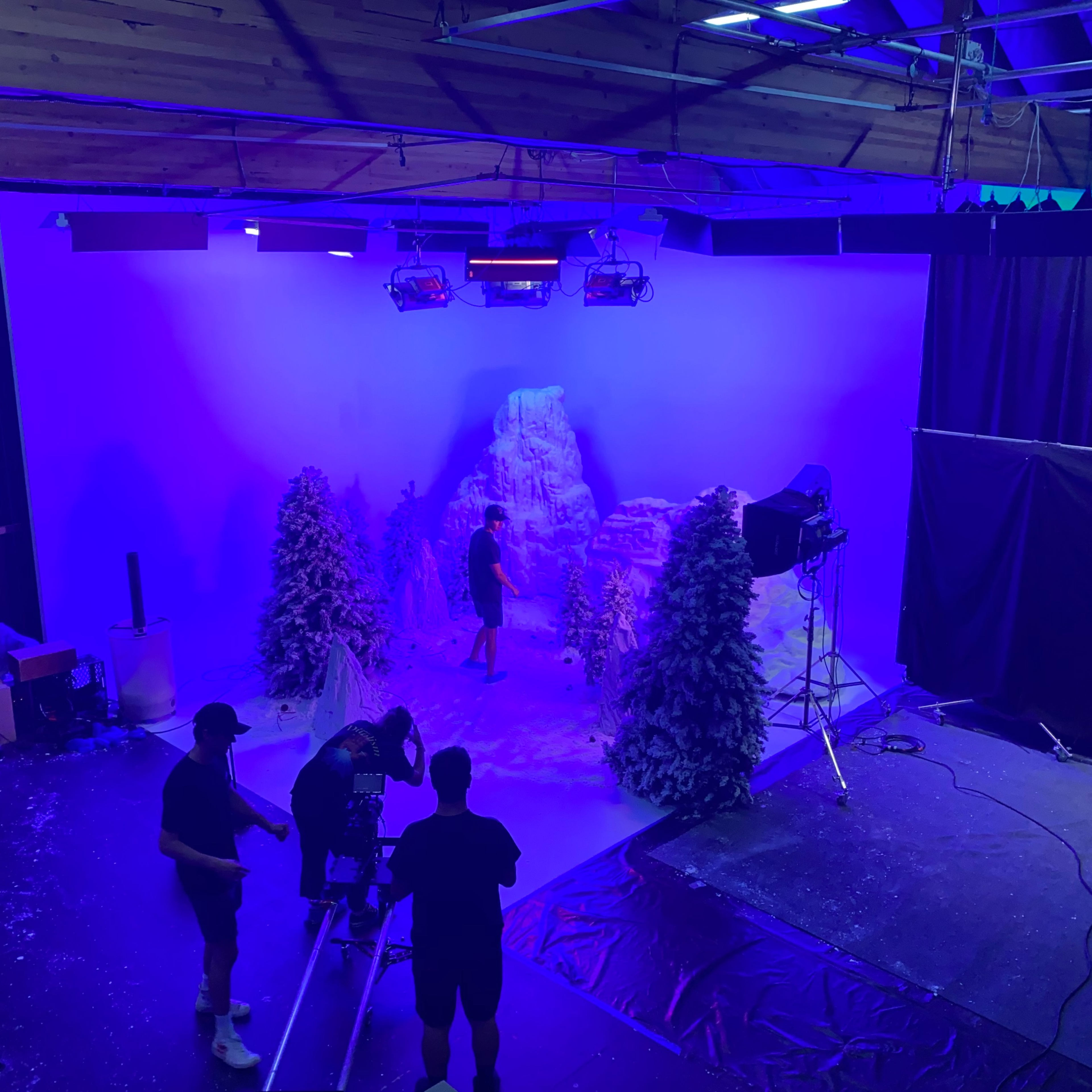 A film crew sets up a winter-themed backdrop featuring artificial snow and pine trees in a studio illuminated by blue lights.