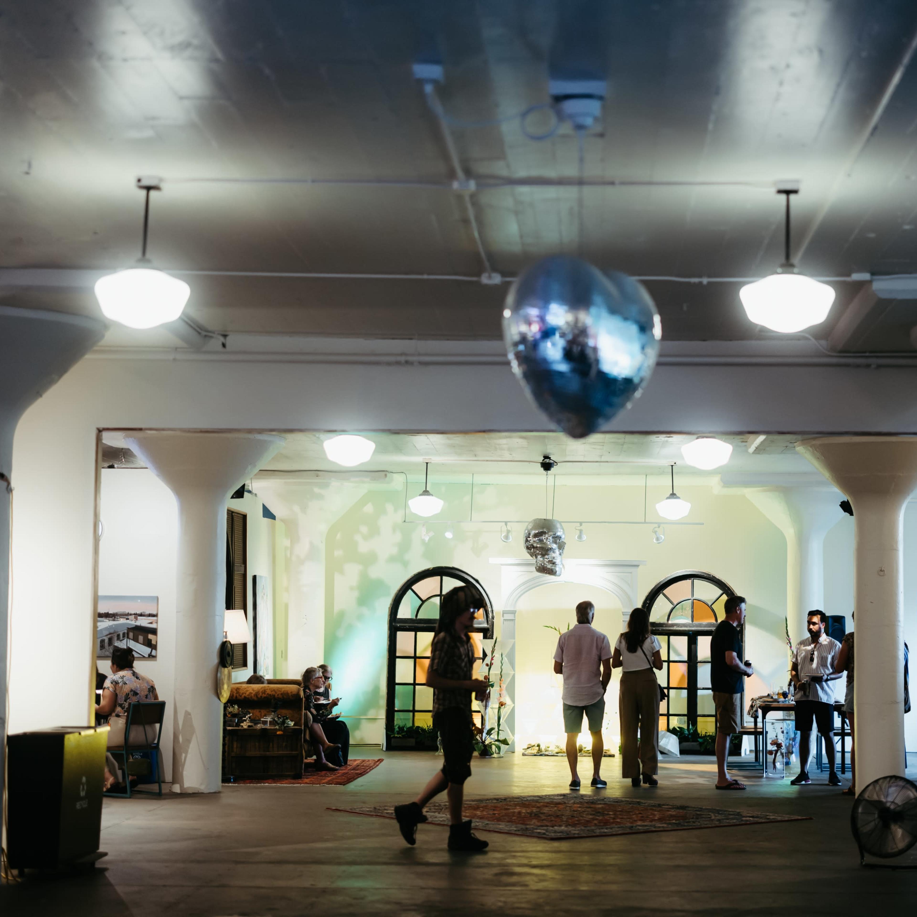 An interior space with polished floors, pillars, and various people engaged in conversation under soft lighting, including a disco ball hanging from the ceiling.
